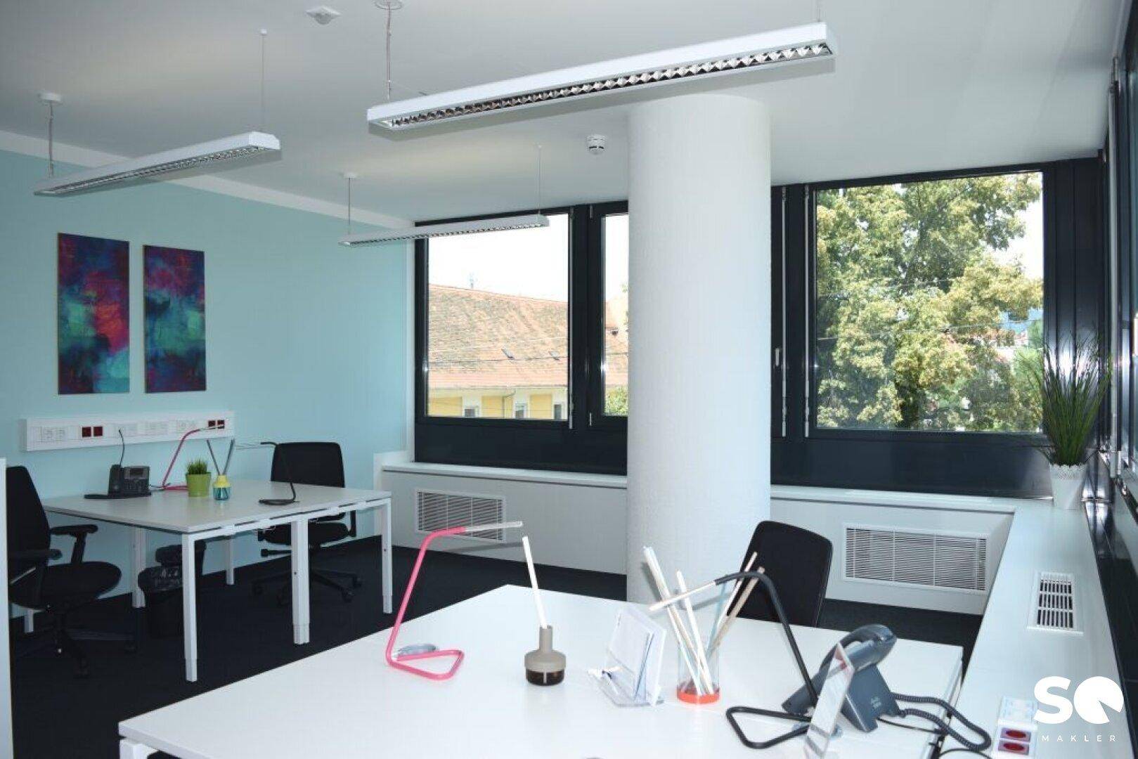 Helles Büro mit zwei Arbeitsplätzen, hellblauen Wänden und großen Fenstern mit Blick ins Grüne.