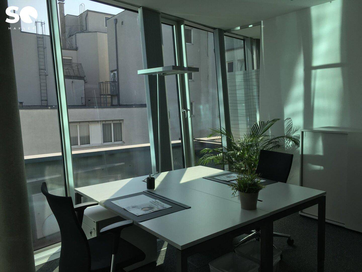 Kleiner Büroraum mit Schreibtisch, Bürostuhl und großen Fenstern mit Blick auf umliegende Gebäude.