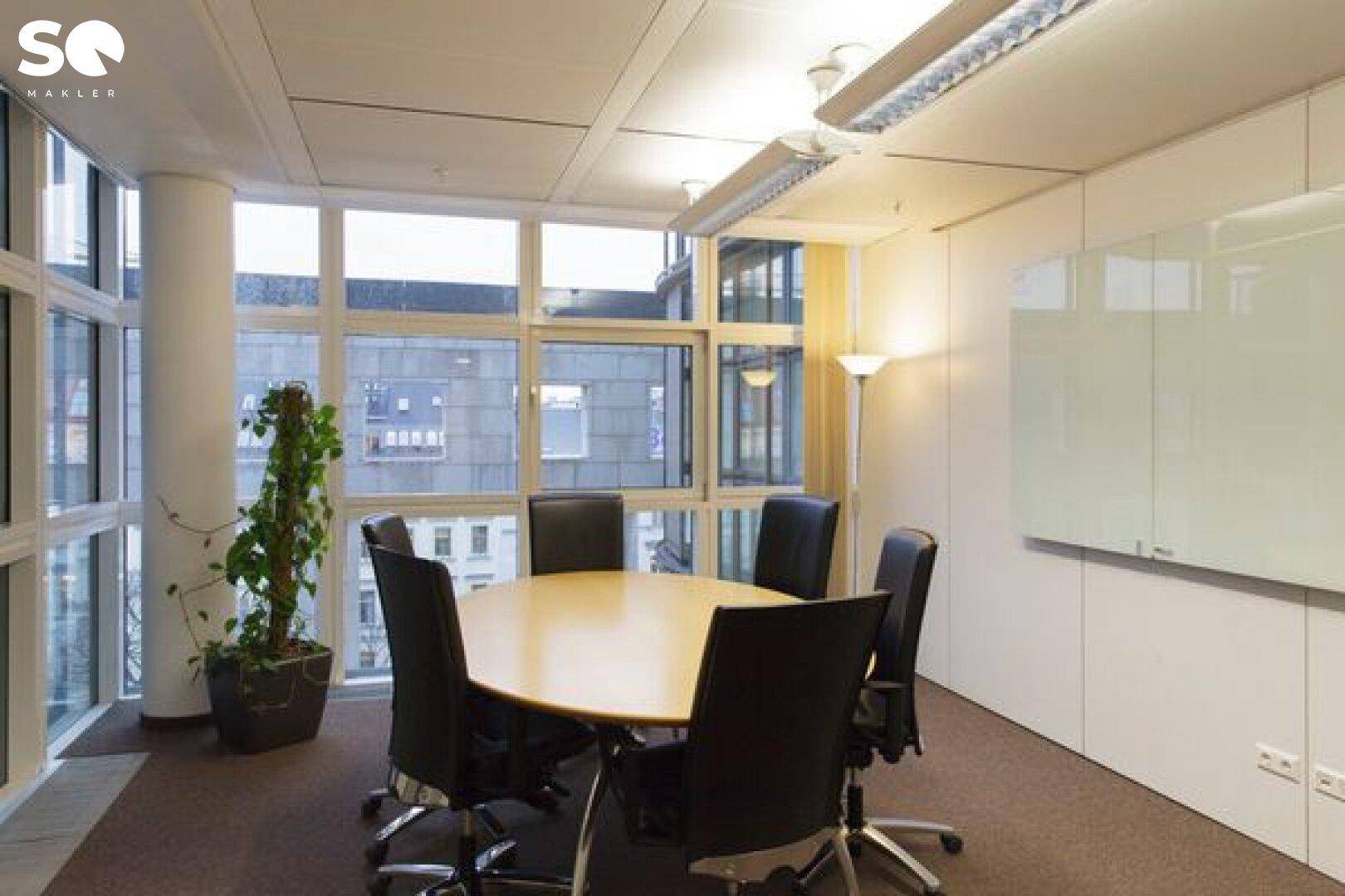 Heller Konferenzraum mit ovalem Holztisch, Ledersesseln und großen Fenstern, die einen weiten Stadtblick bieten.