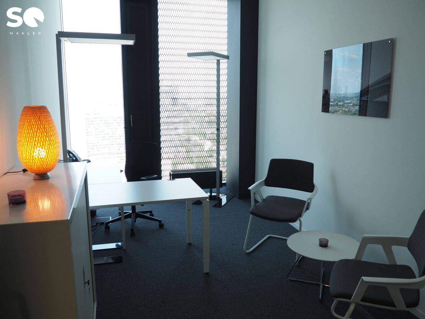 Einzelbüro mit Schreibtisch, bequemen Stühlen und dekorativer Beleuchtung neben einem großen Fenster.