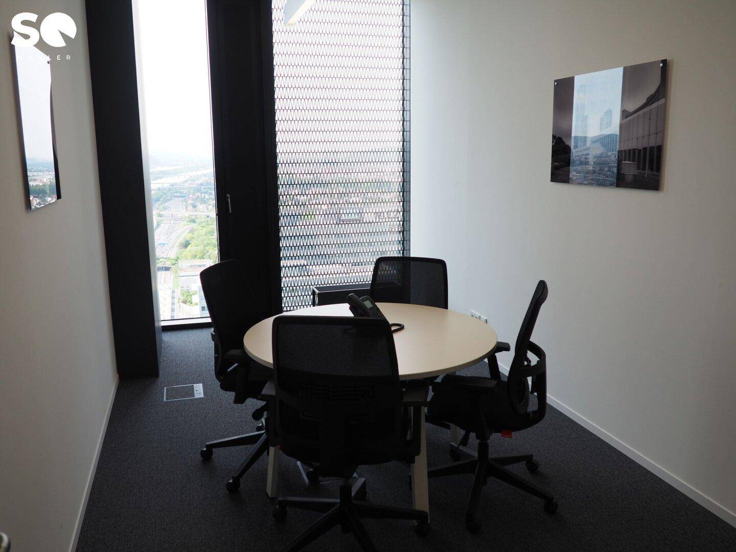 Kleiner Besprechungsraum mit rundem Tisch, Bürostühlen und großem Fenster mit Stadtblick.