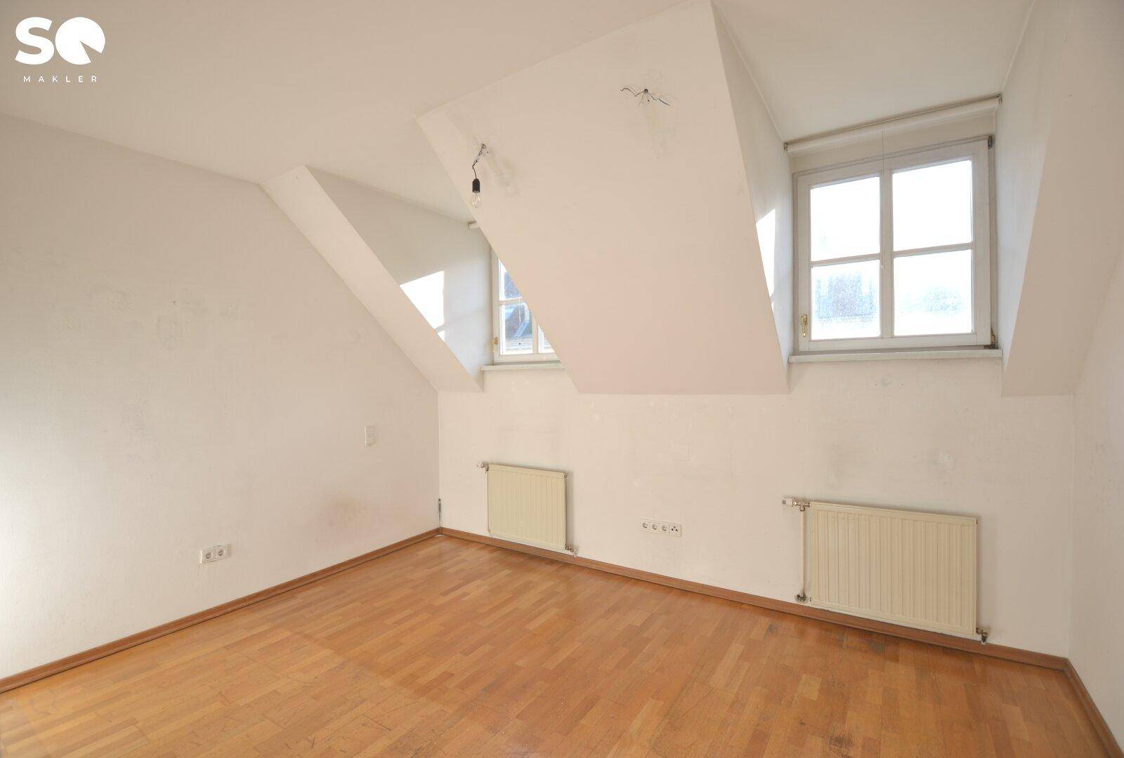 Schlafzimmer mit Holzboden, Dachschrägen und zwei Fenstern für natürliche Belichtung.