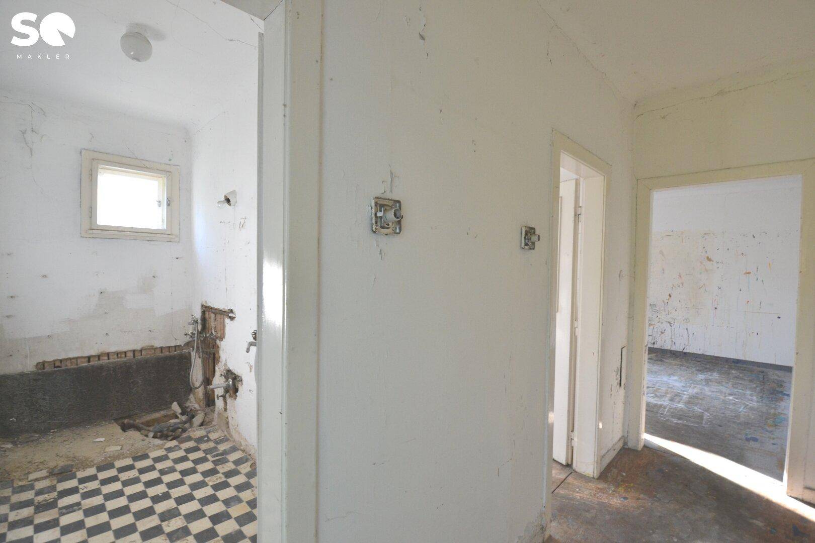 Flur mit Zugang zu einem Badezimmer in Renovierung, mit karierten Fliesen und freiliegenden Leitungen.