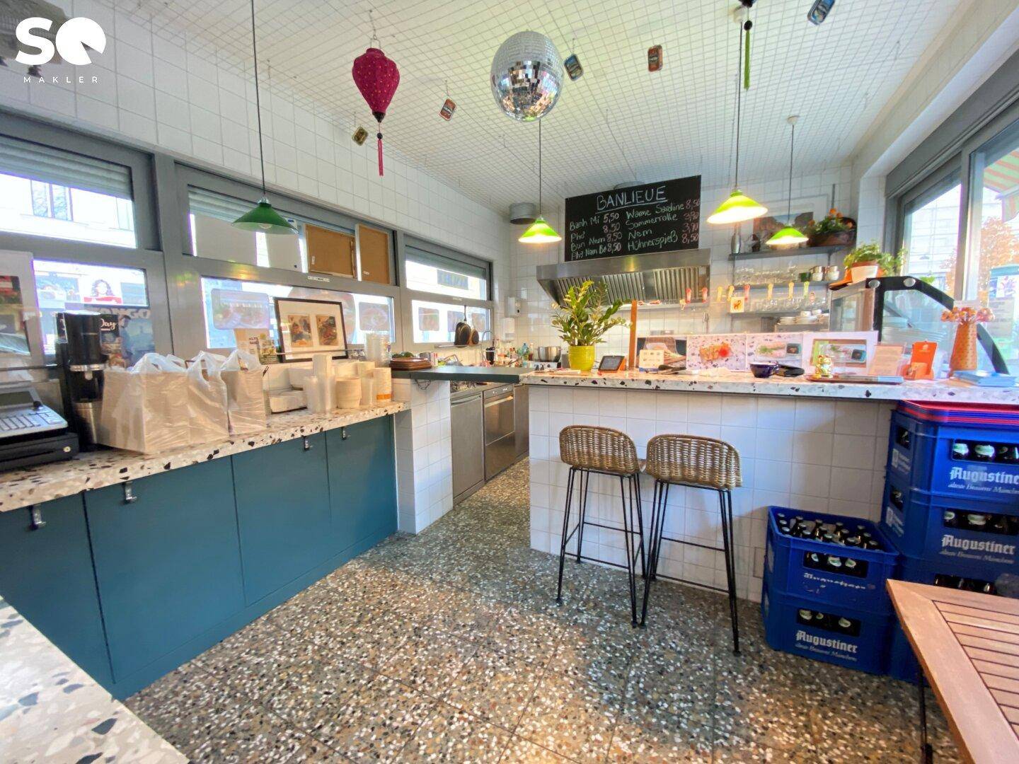 Innenansicht eines kleinen Restaurants mit Theke, Barhockern und Fliesenboden, hell durch große Fenster.