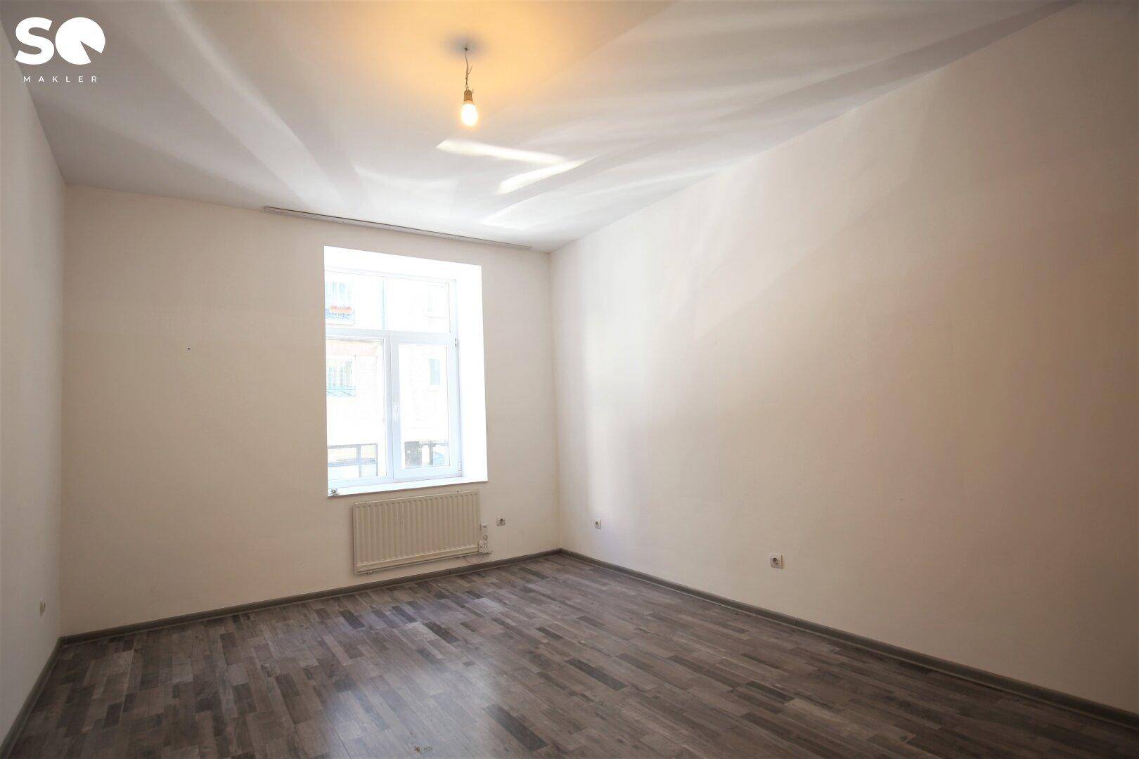 Geräumiger, heller Raum mit Laminatboden, einem Fenster und einem Heizkörper darunter.