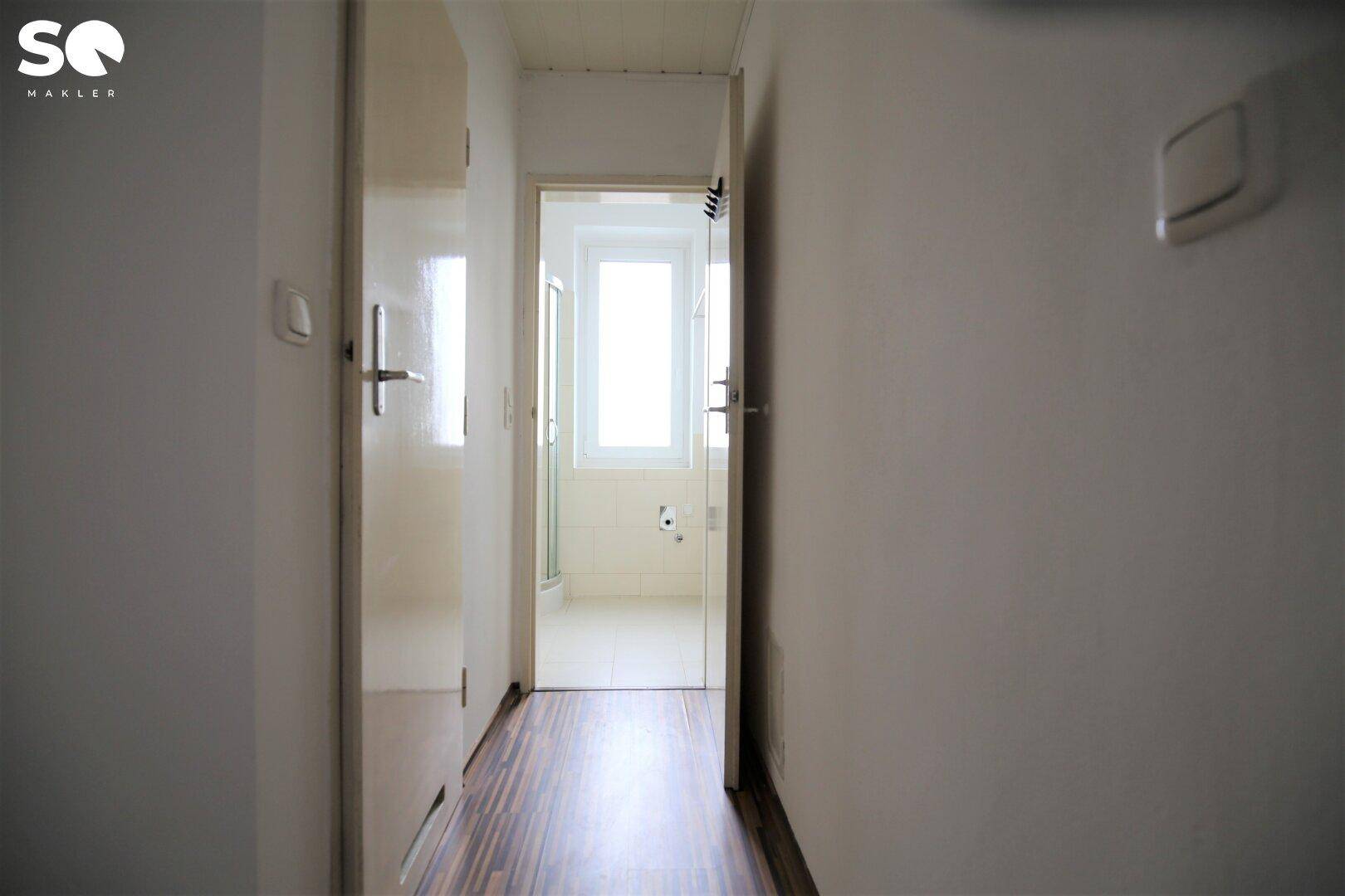 Flur mit Laminatboden und Blick in ein helles Badezimmer mit Fenster.