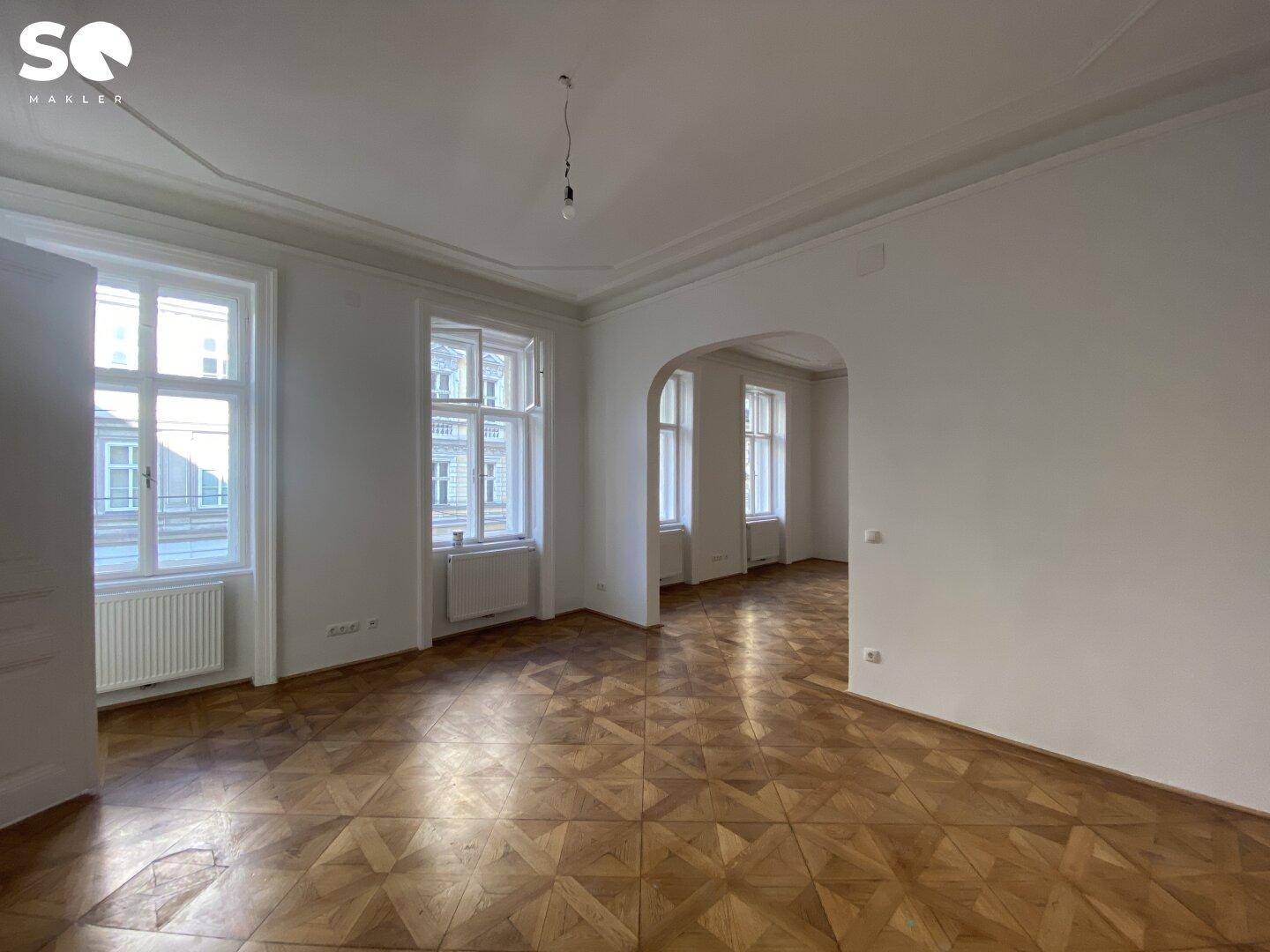 Geräumiger Wohnbereich mit hellem Fischgrätparkett, hohen Decken und einem eleganten Bogen zu einem weiteren Zimmer.