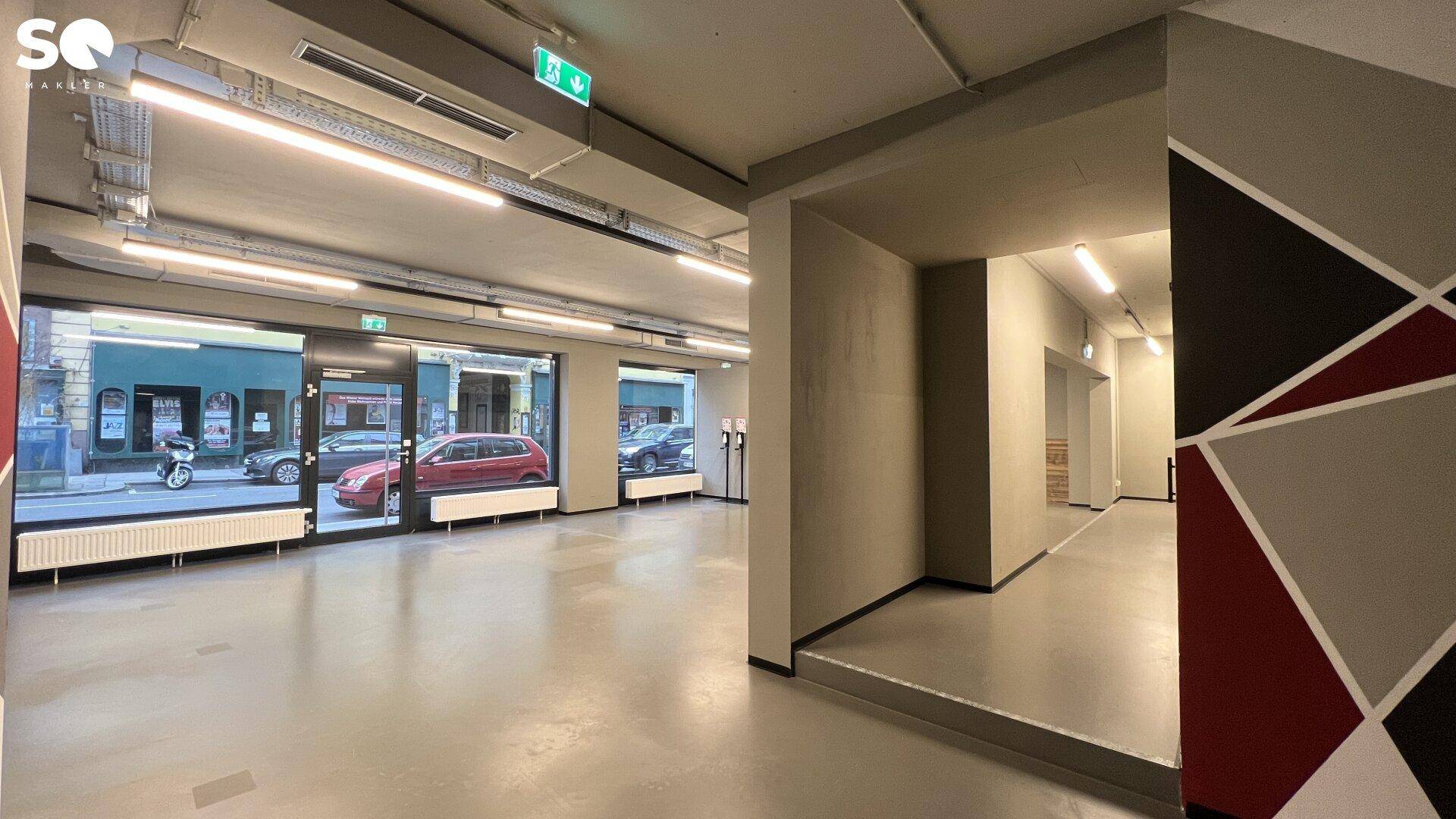 Geräumiger Gewerberaum mit großen Schaufenstern, sichtbaren Deckeninstallationen und einem modernen geometrischen Wandmuster.