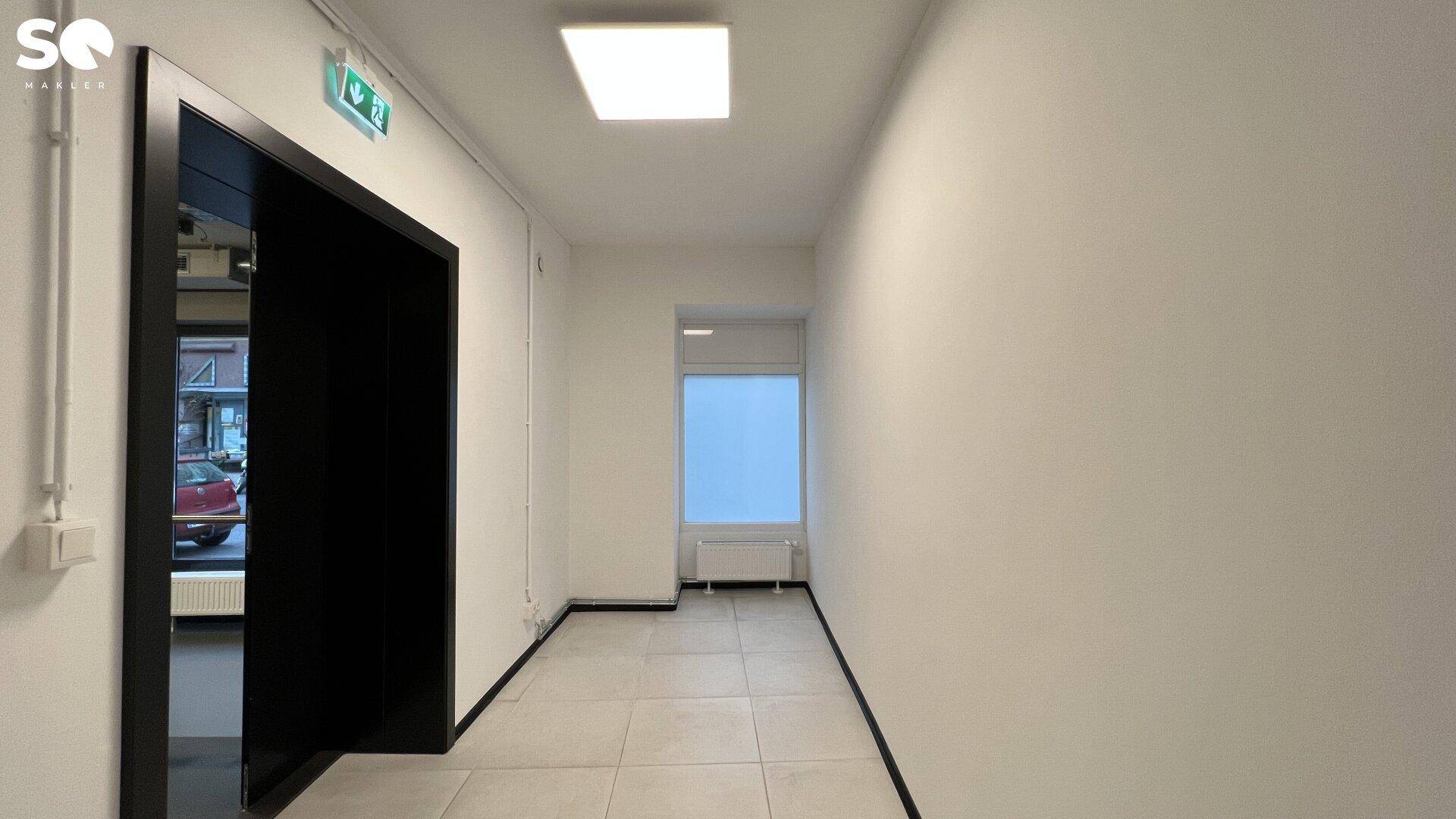 Heller Flur mit hellen Fliesen, einem Fenster am Ende und einem dunklen Türrahmen auf der linken Seite.