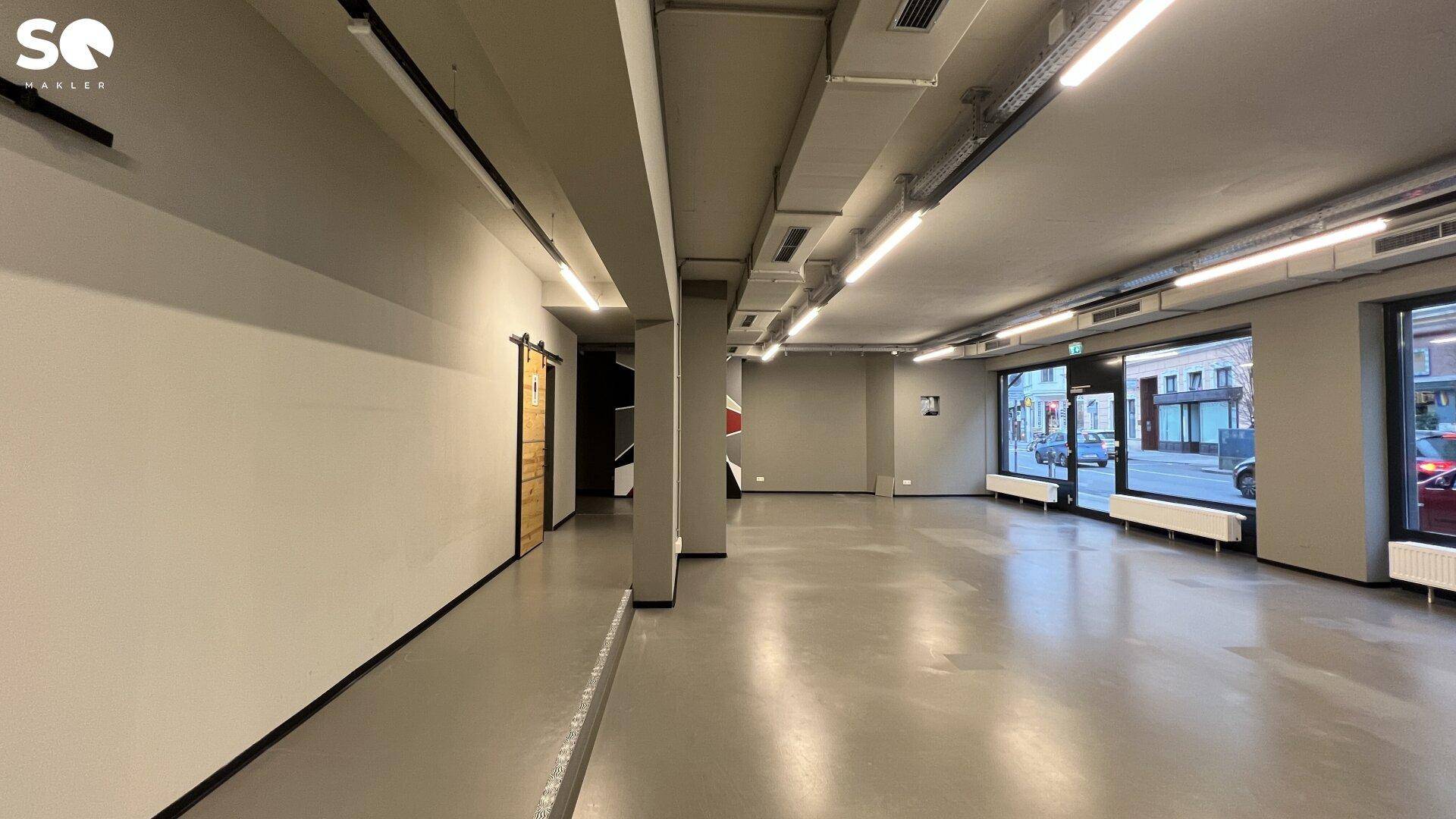 Langer, offener Gewerberaum mit großen Fenstern, sichtbaren Deckenleitungen und einer modernen Holzschiebetür.