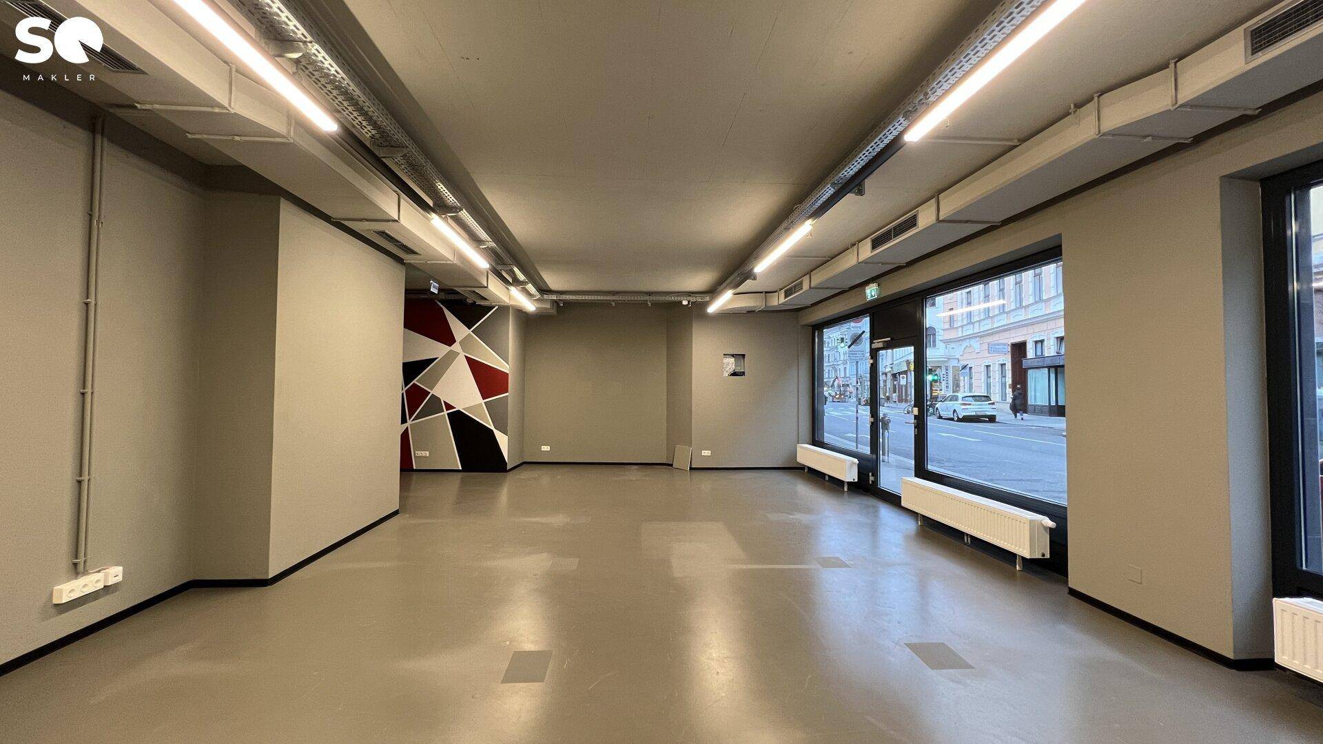 Weitläufiger Gewerberaum mit viel Tageslicht, sichtbaren Deckeninstallationen und einer auffälligen geometrischen Wandgestaltung.
