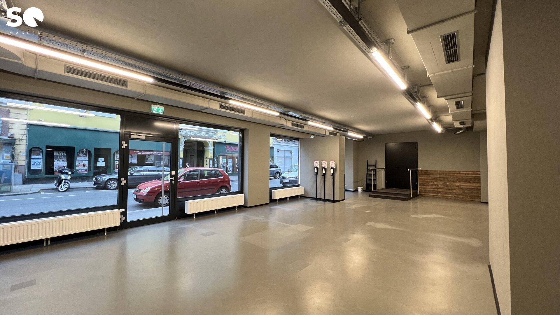 Heller Gewerberaum mit großen Schaufenstern, industrieller Deckenästhetik und einer kleinen erhöhten Plattform.