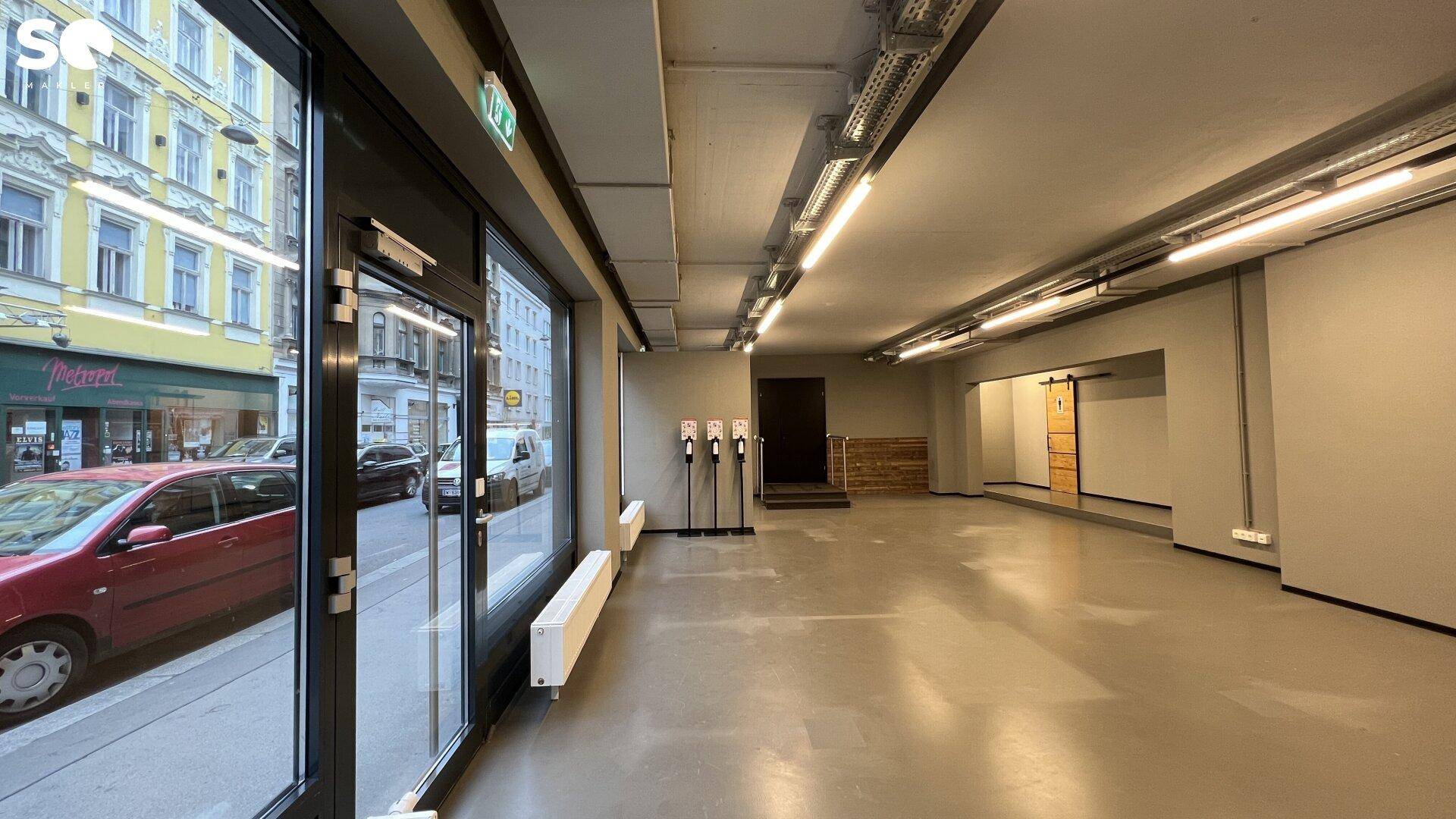 Geräumiger Gewerberaum mit großen Schaufenstern, sichtbaren Deckeninstallationen und viel Tageslicht von der Straße.