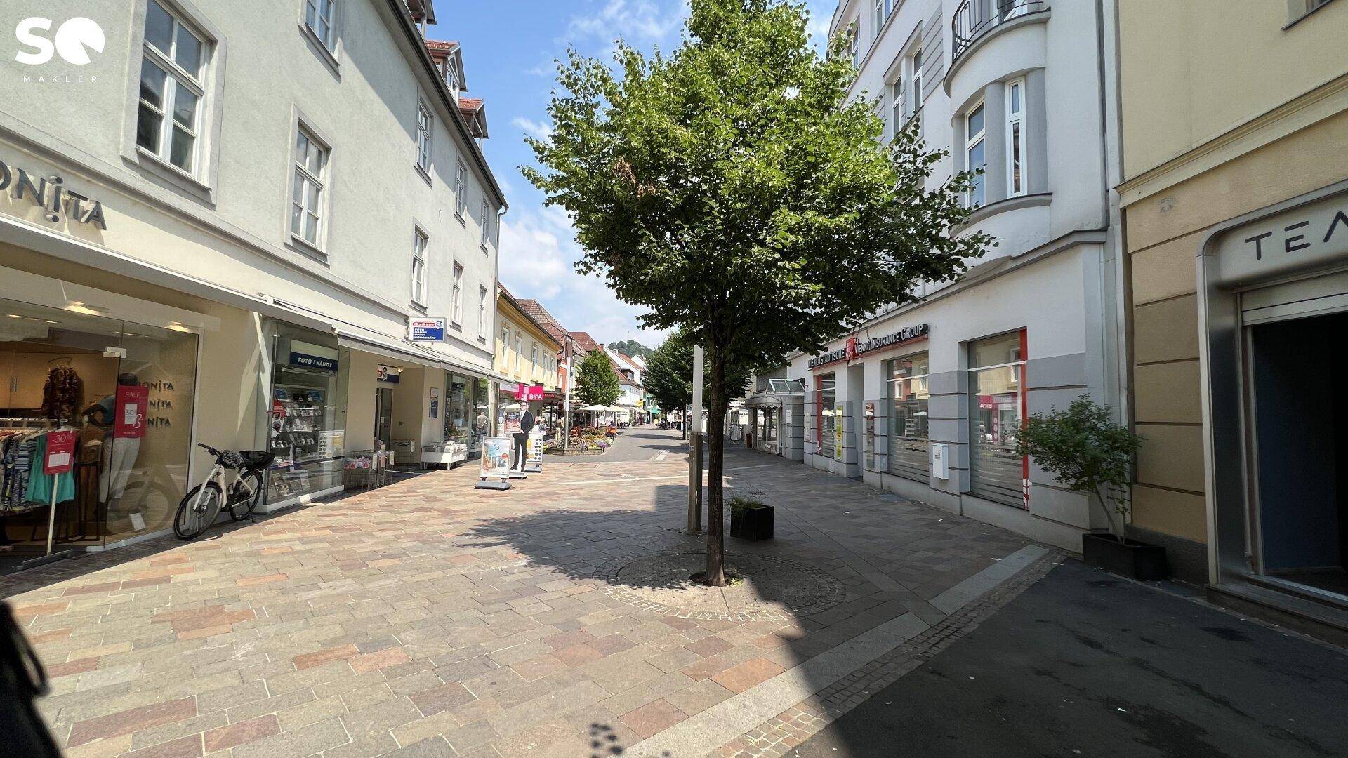 Eine belebte Fußgängerzone mit Geschäften und Bäumen, umgeben von historischen Stadtgebäuden.