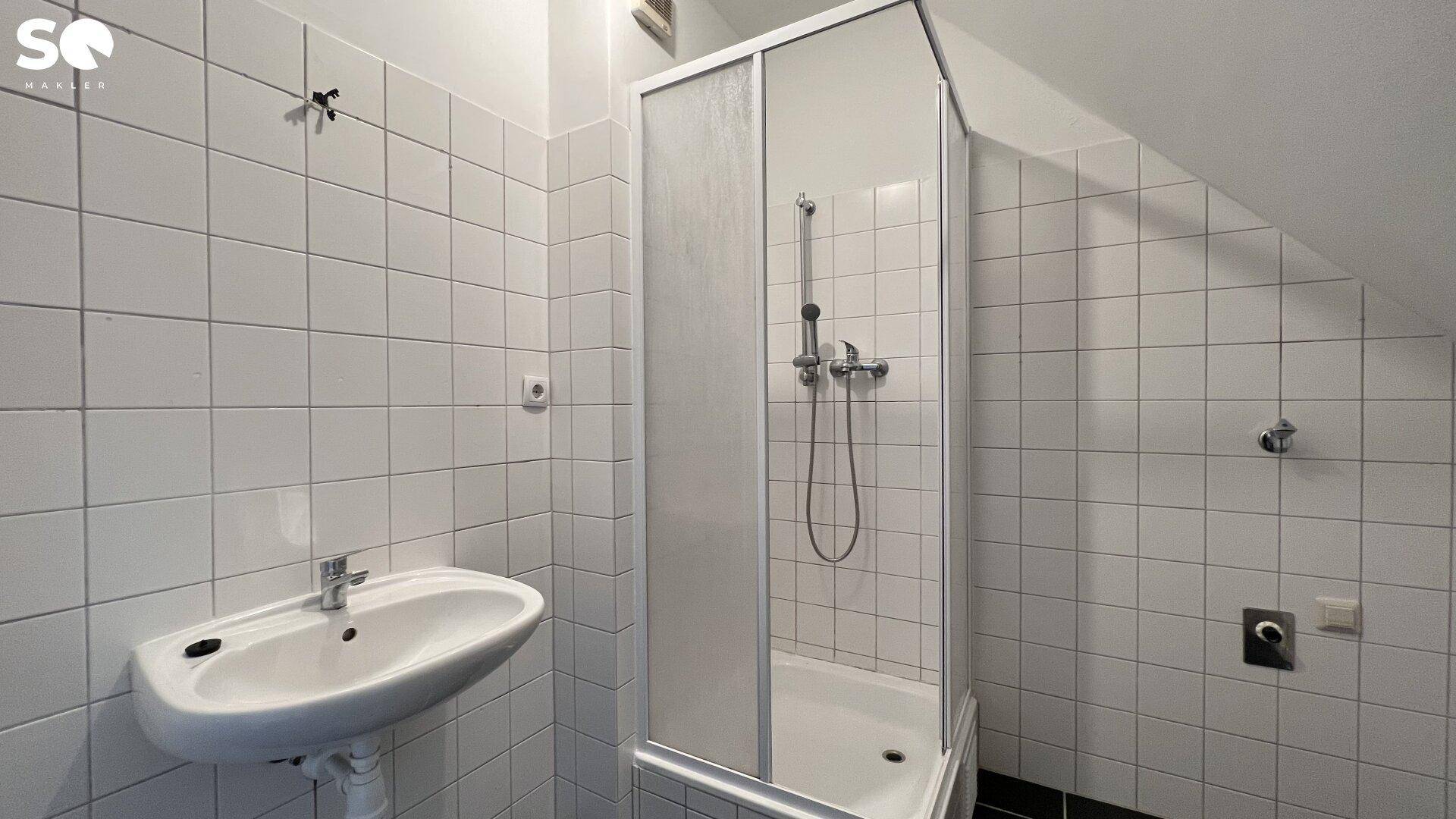Funktionales Badezimmer mit Duschkabine, Waschbecken und weiß-schwarzen Fliesen unter der Dachschräge.