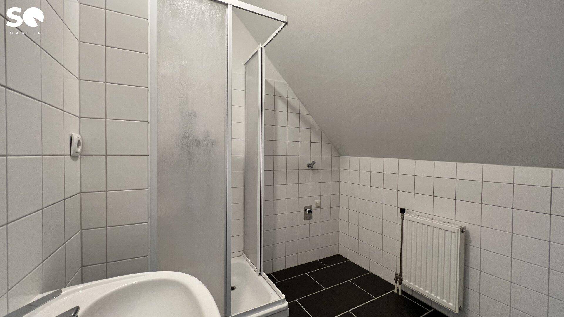 Badezimmer mit weiß gefliesten Wänden, schwarzem Boden, Duschkabine und Dachschräge.