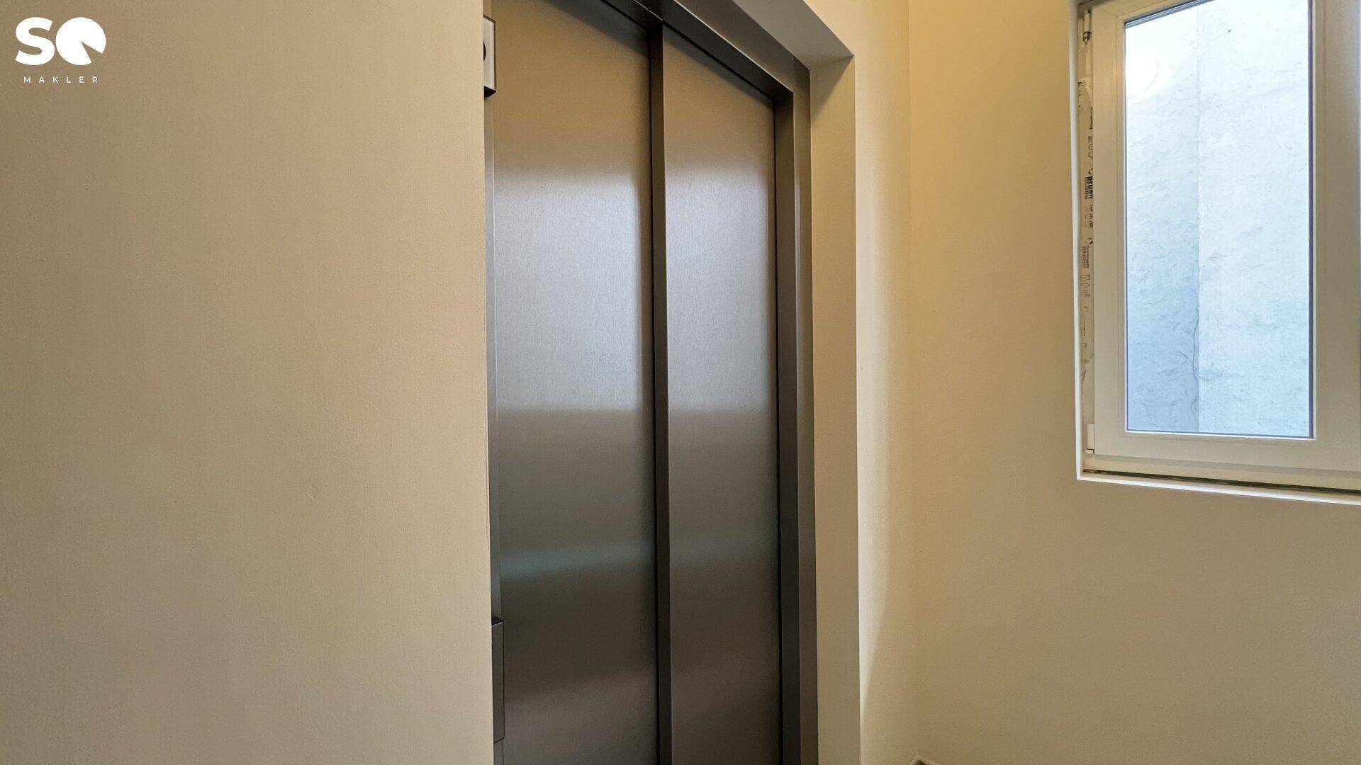 Moderne Aufzugstüren aus Edelstahl neben einem Fenster im Treppenhaus.