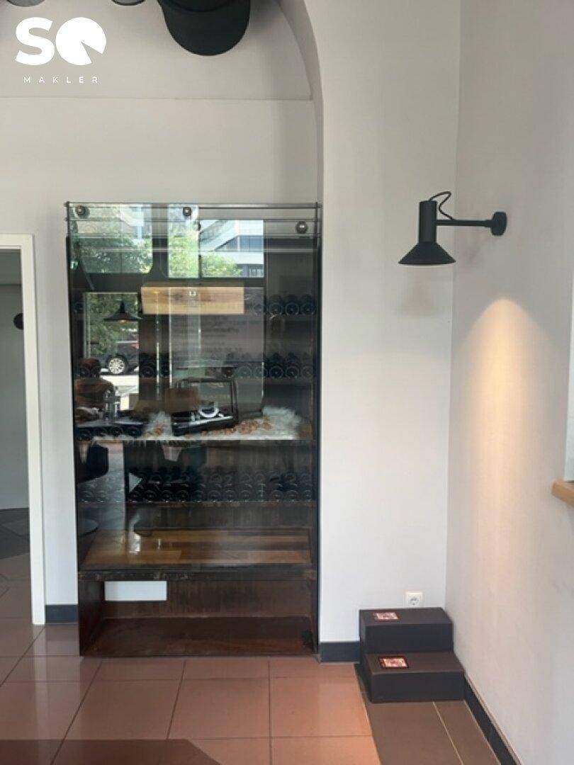 Elegante Glasvitrine präsentiert Produkte, beleuchtet von einer modernen Wandlampe, auf einem hellen Fliesenboden.