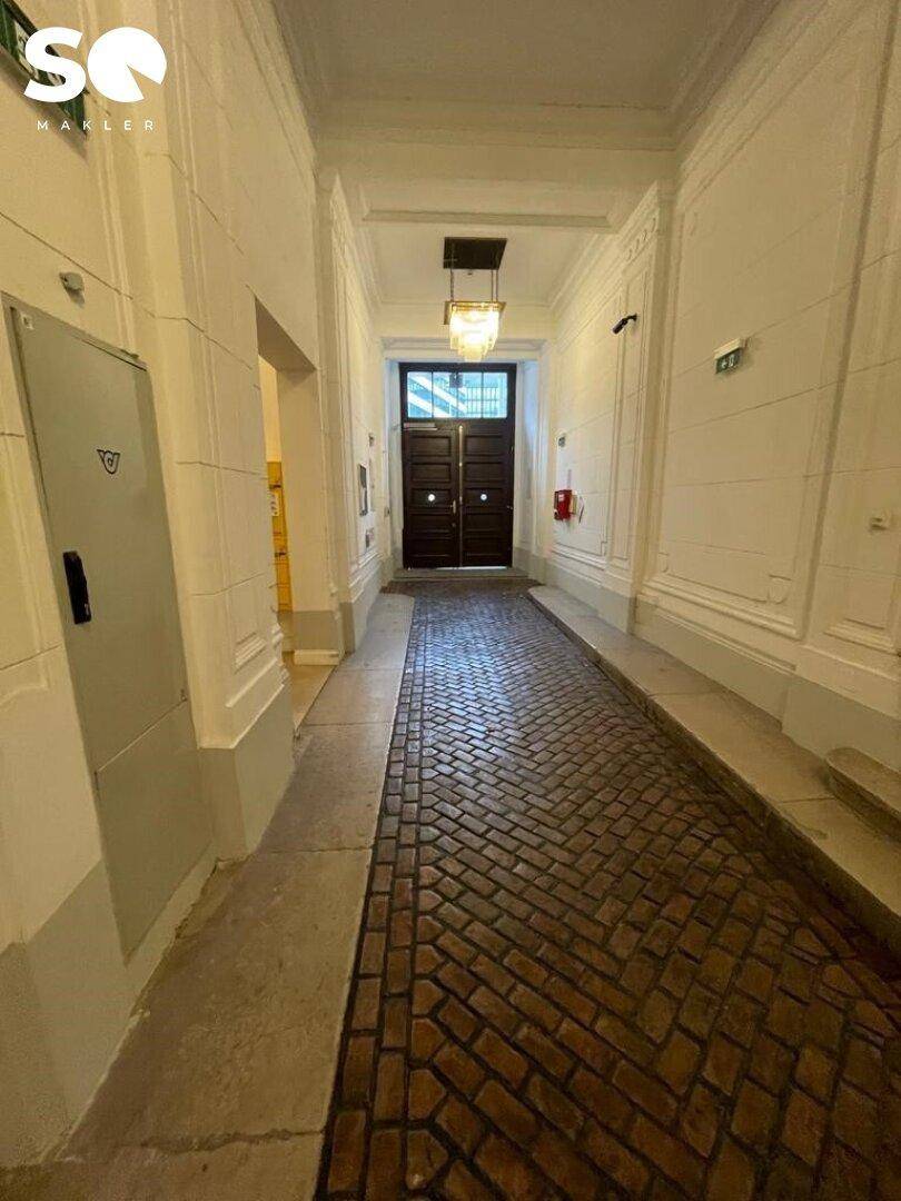 Historischer Eingangsbereich mit Ziegelmusterboden und einer eleganten Deckenleuchte, die zum Gebäude führt.