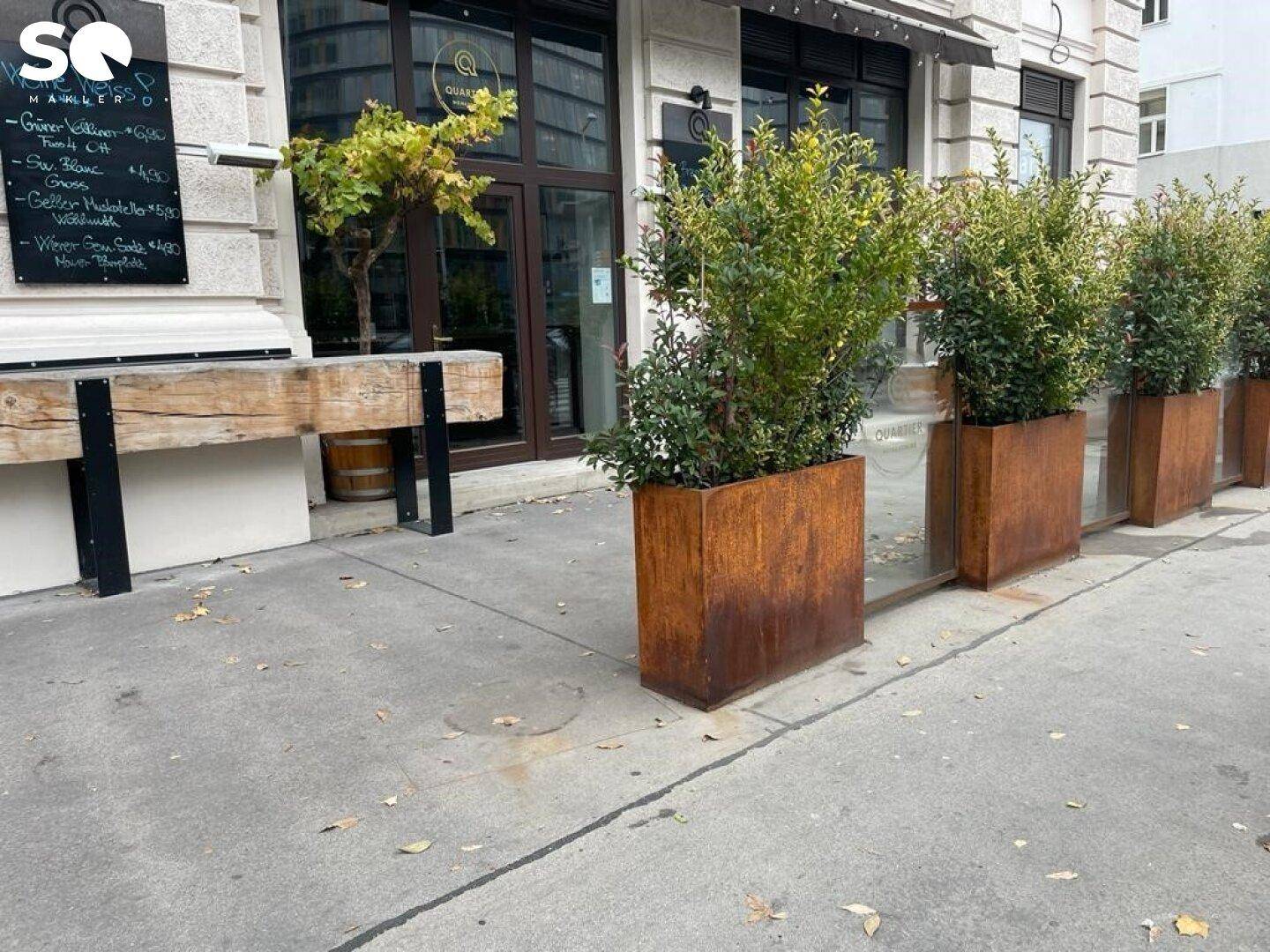 Nahaufnahme des Geschäftseingangs mit rostfarbenen Pflanzenkübeln und einer Kreidetafel mit Tagesangeboten.