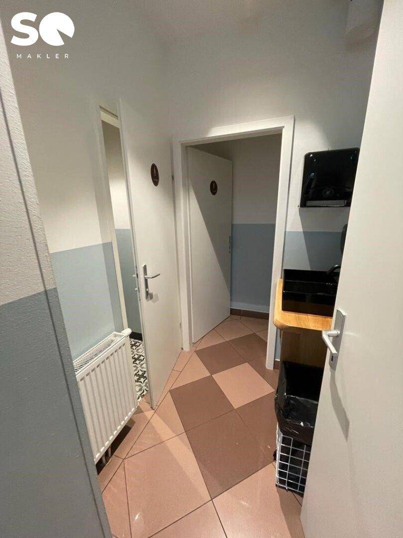 Flur mit gemusterten Fliesen, der zu den Toiletten führt, und einem Heizkörper an der Wand.