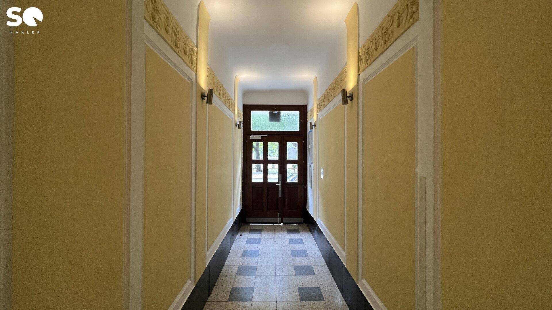 Langer Flur in einem Altbau mit gelben Wänden, gemustertem Fliesenboden und Holztür am Ende.
