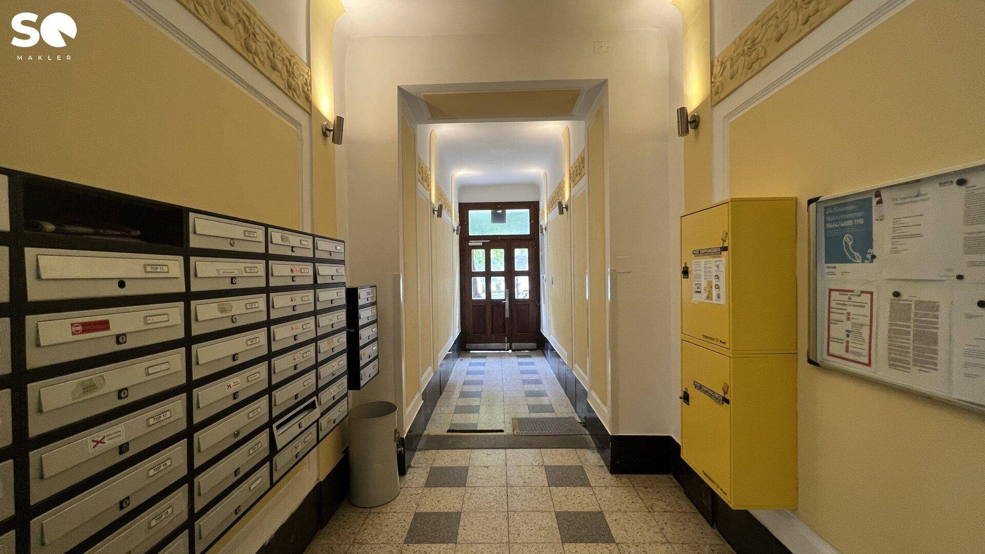 Eingangsbereich eines Gebäudes mit Briefkästen, Informationswand und gefliestem Boden.