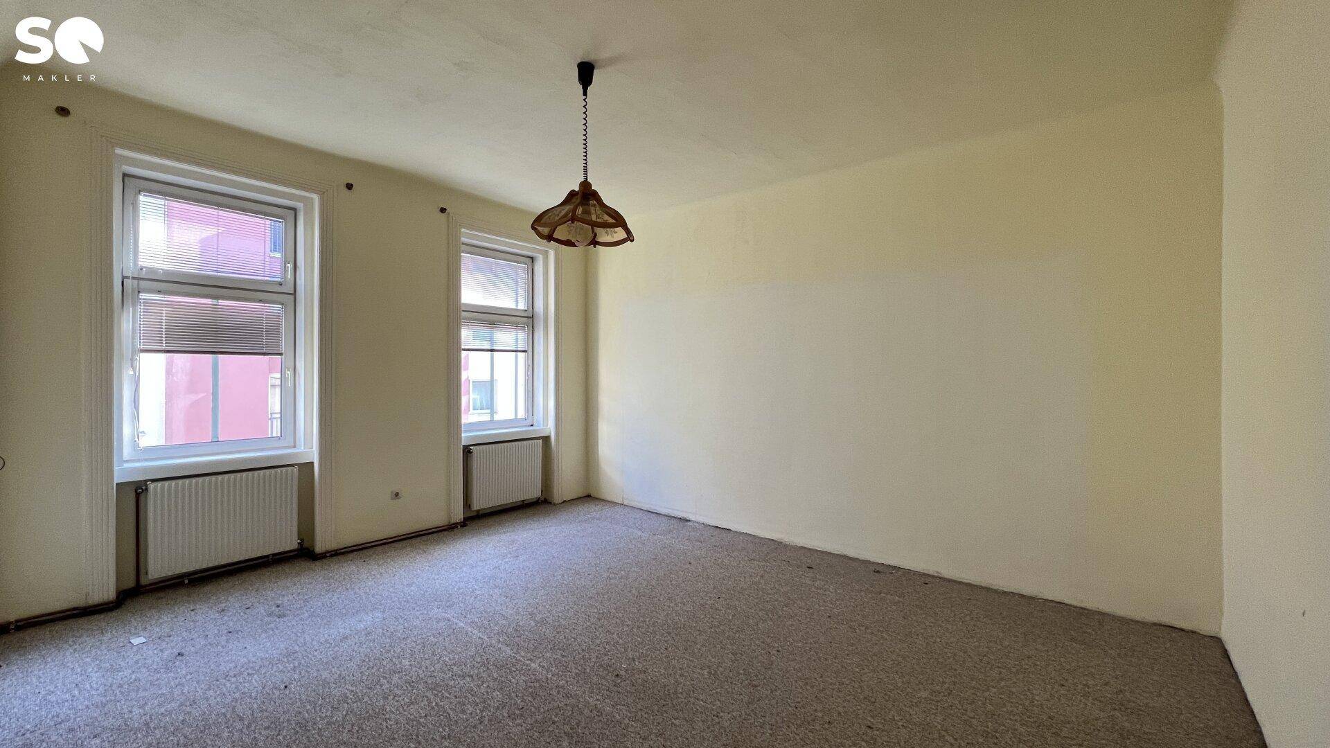 Geräumiges Zimmer mit Teppichboden, zwei Fenstern und einer einfachen Deckenleuchte.