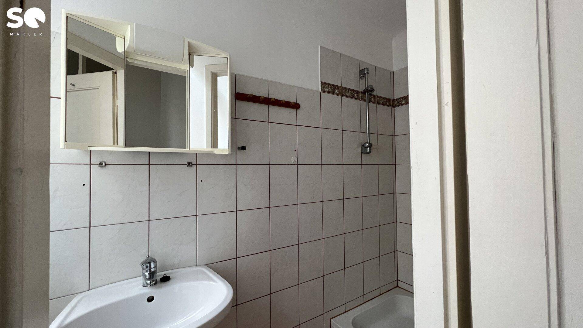Badezimmer mit weißen Fliesen, Waschbecken und Spiegelschrank, renovierungsbedürftig.