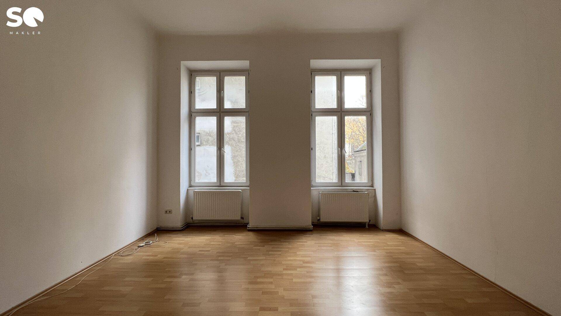 Helles, leeres Zimmer mit Holzboden und zwei großen Fenstern, bietet viel Tageslicht.