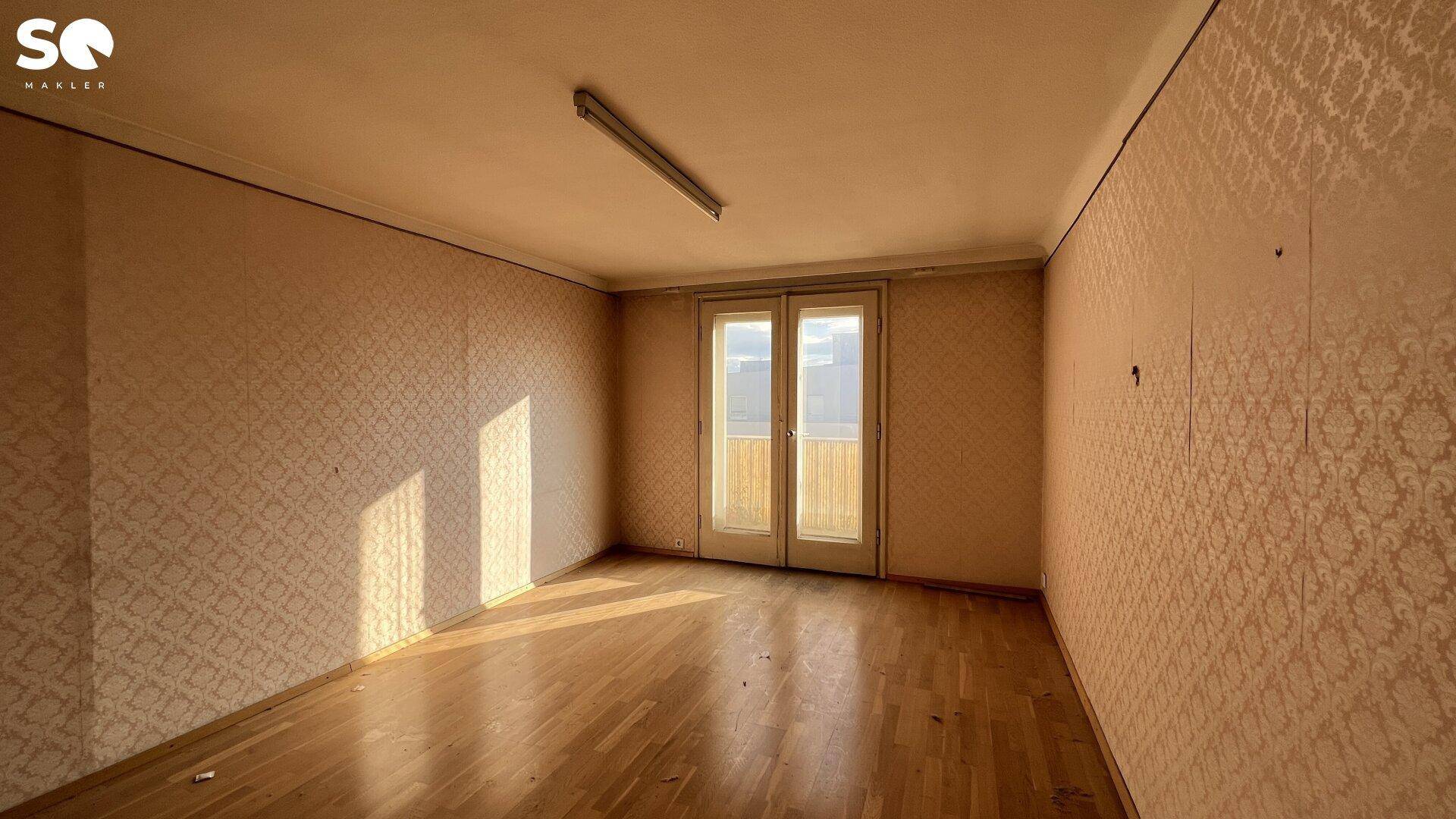 Unmöbliertes Zimmer mit gemusterter Tapete, Holzboden und Balkonzugang.