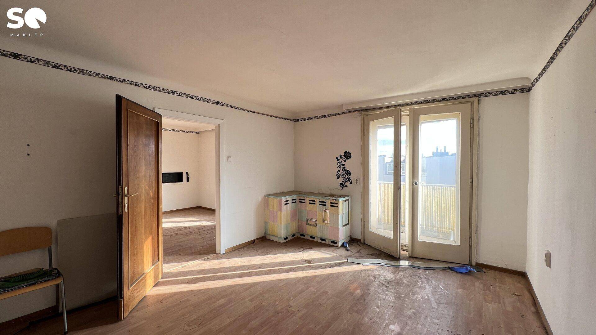 Unmöbliertes Wohnzimmer mit Holzboden, Balkonzugang und Renovierungsbedarf.