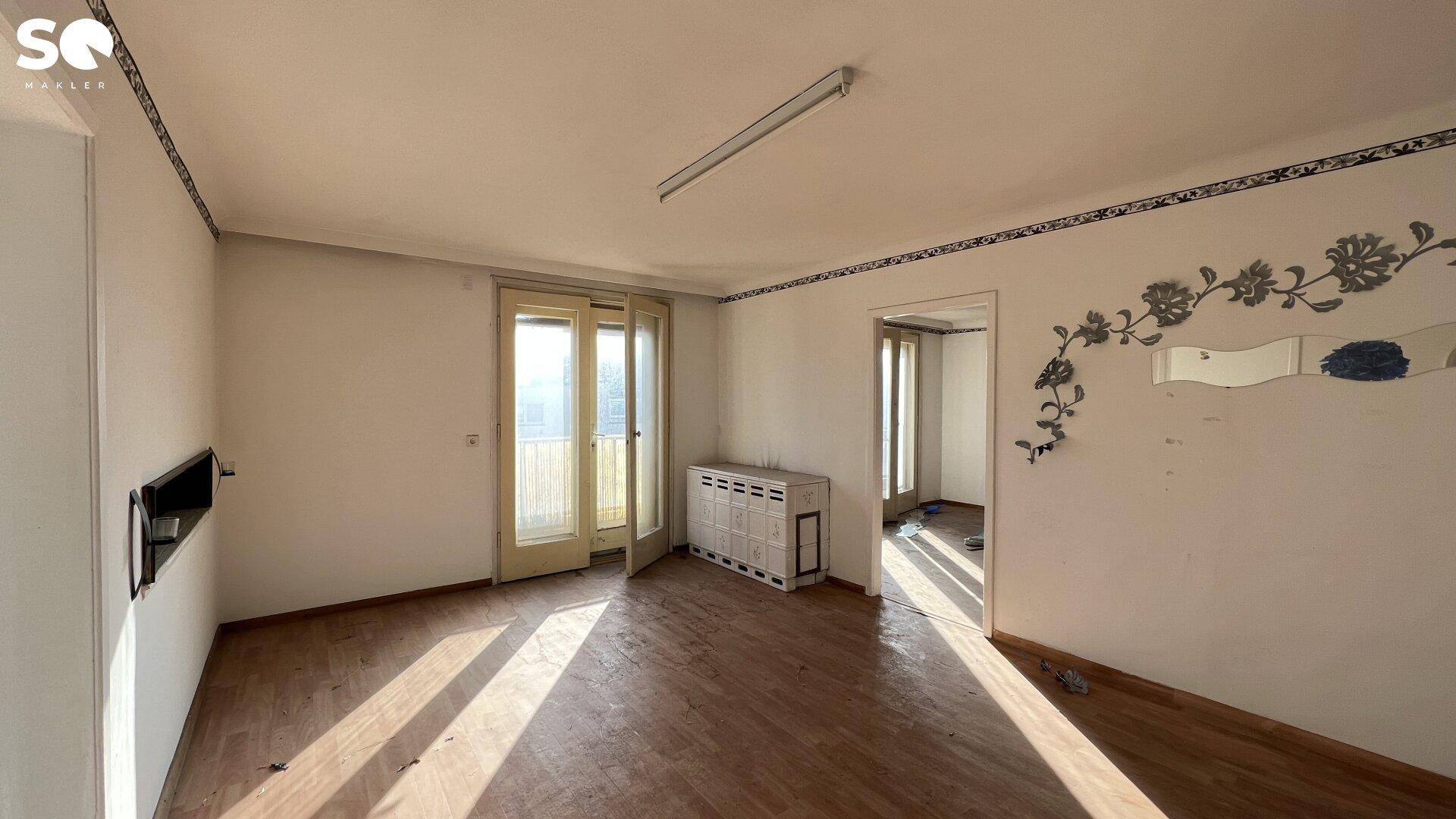Unmöbliertes Wohnzimmer mit Holzboden, Balkonzugang und Renovierungsbedarf.