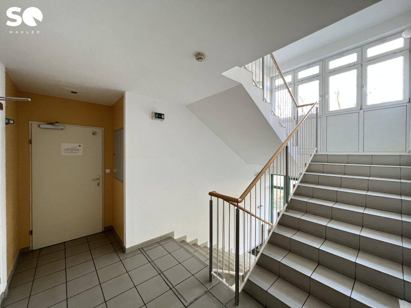 Helles Treppenhaus mit großen Fenstern, Holzgeländer und gefliestem Boden, das zu oberen Etagen führt.