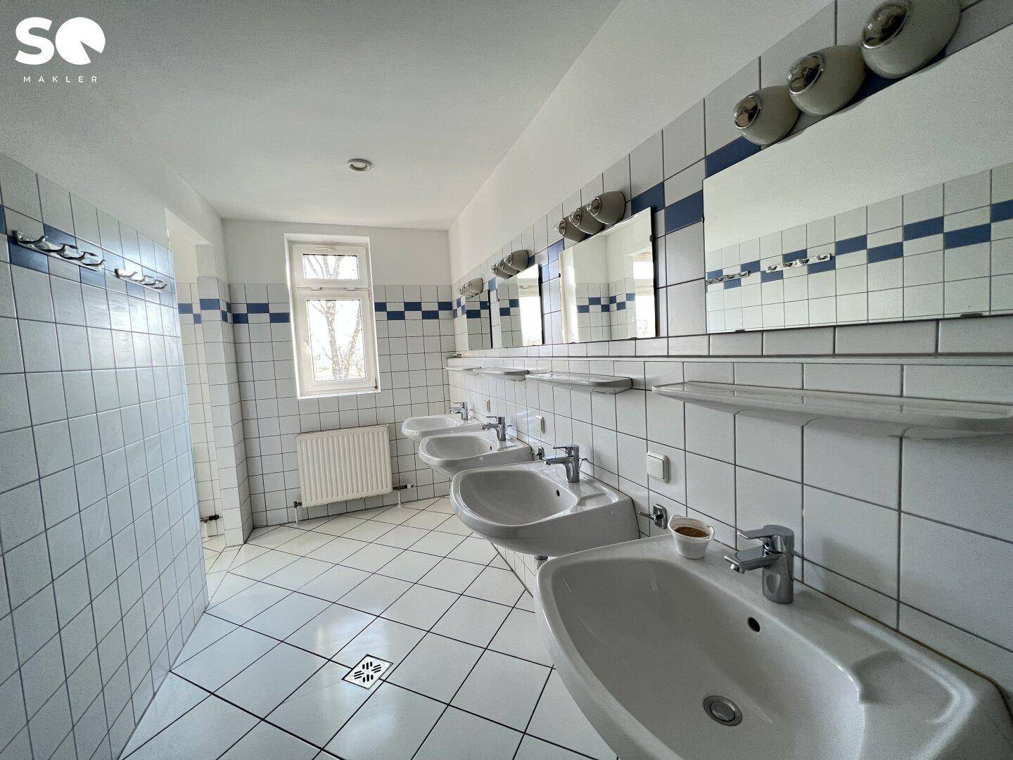 Badezimmer mit mehreren Waschbecken, weißen und blauen Fliesen sowie einem Fenster für Tageslicht.