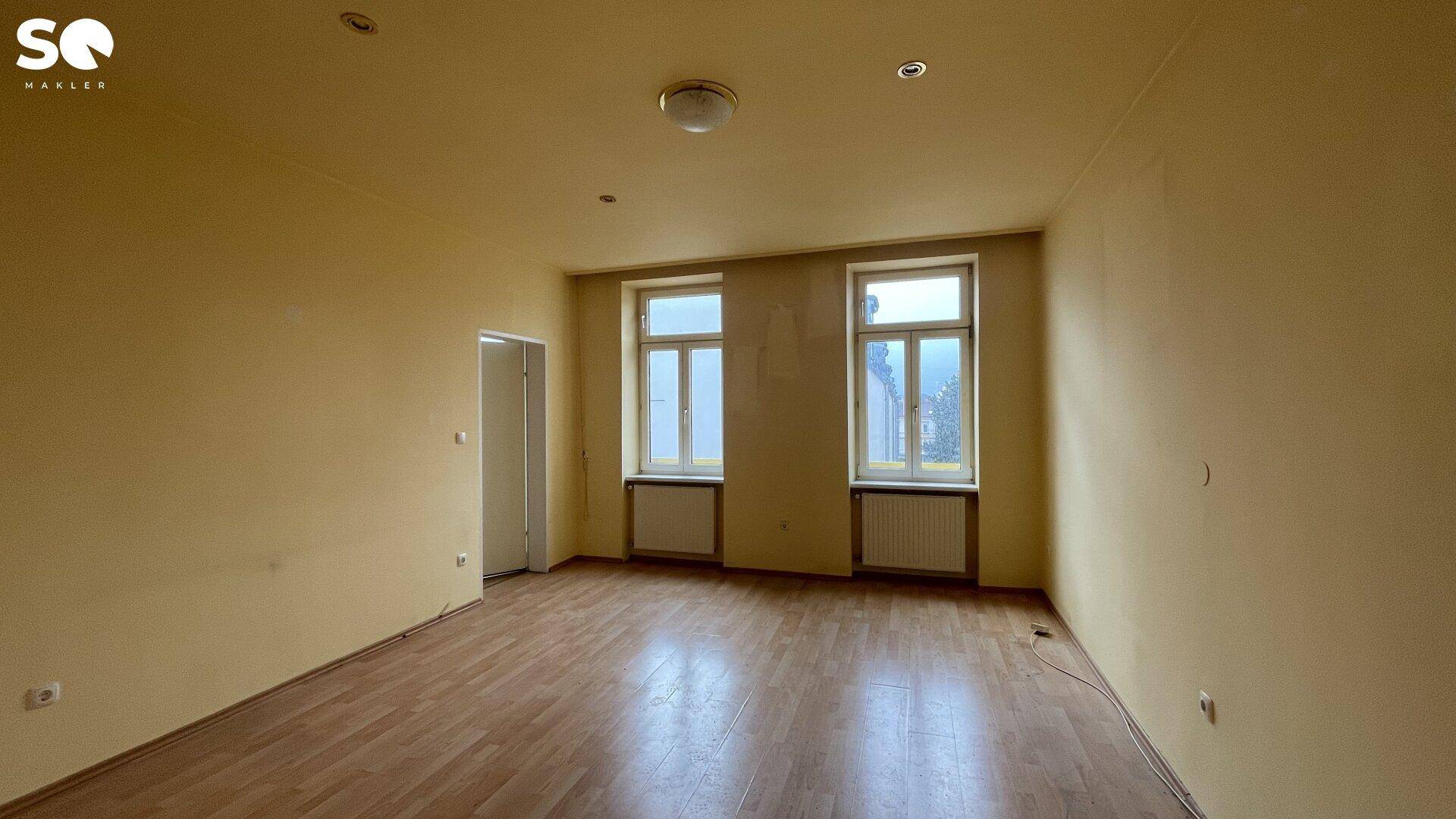 Helles Zimmer mit gelblichen Wänden, Holzboden und zwei Fenstern für viel Licht.