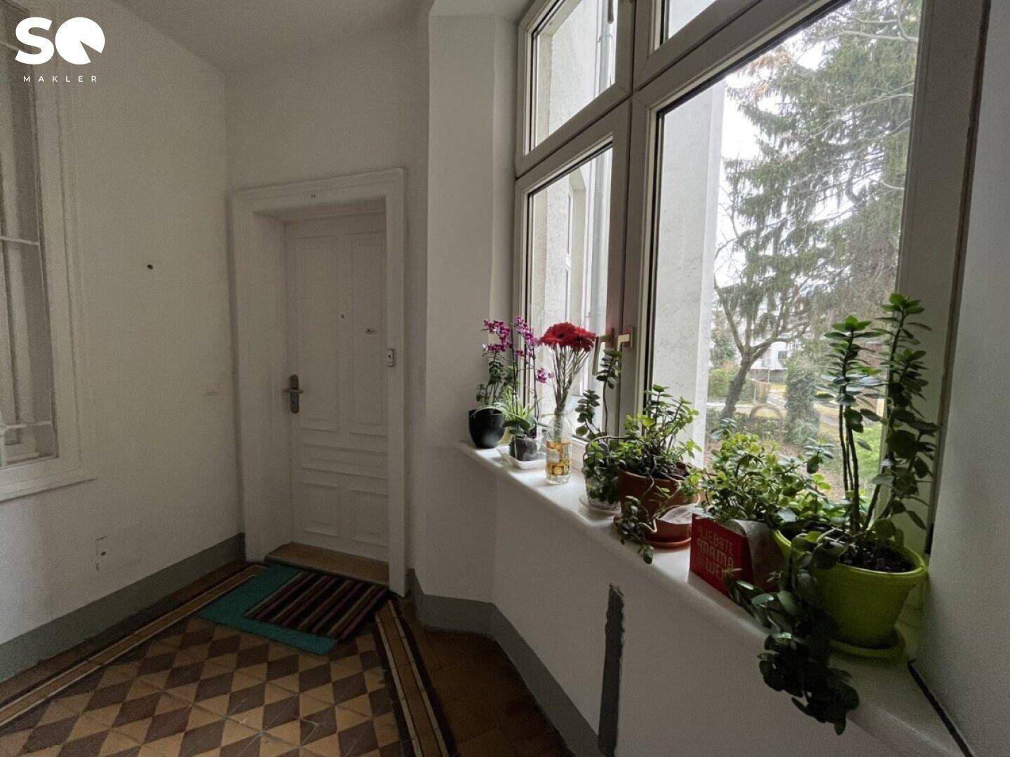 Heller Flur mit großen Fenstern, Pflanzen auf der Fensterbank und gemustertem Fliesenboden.