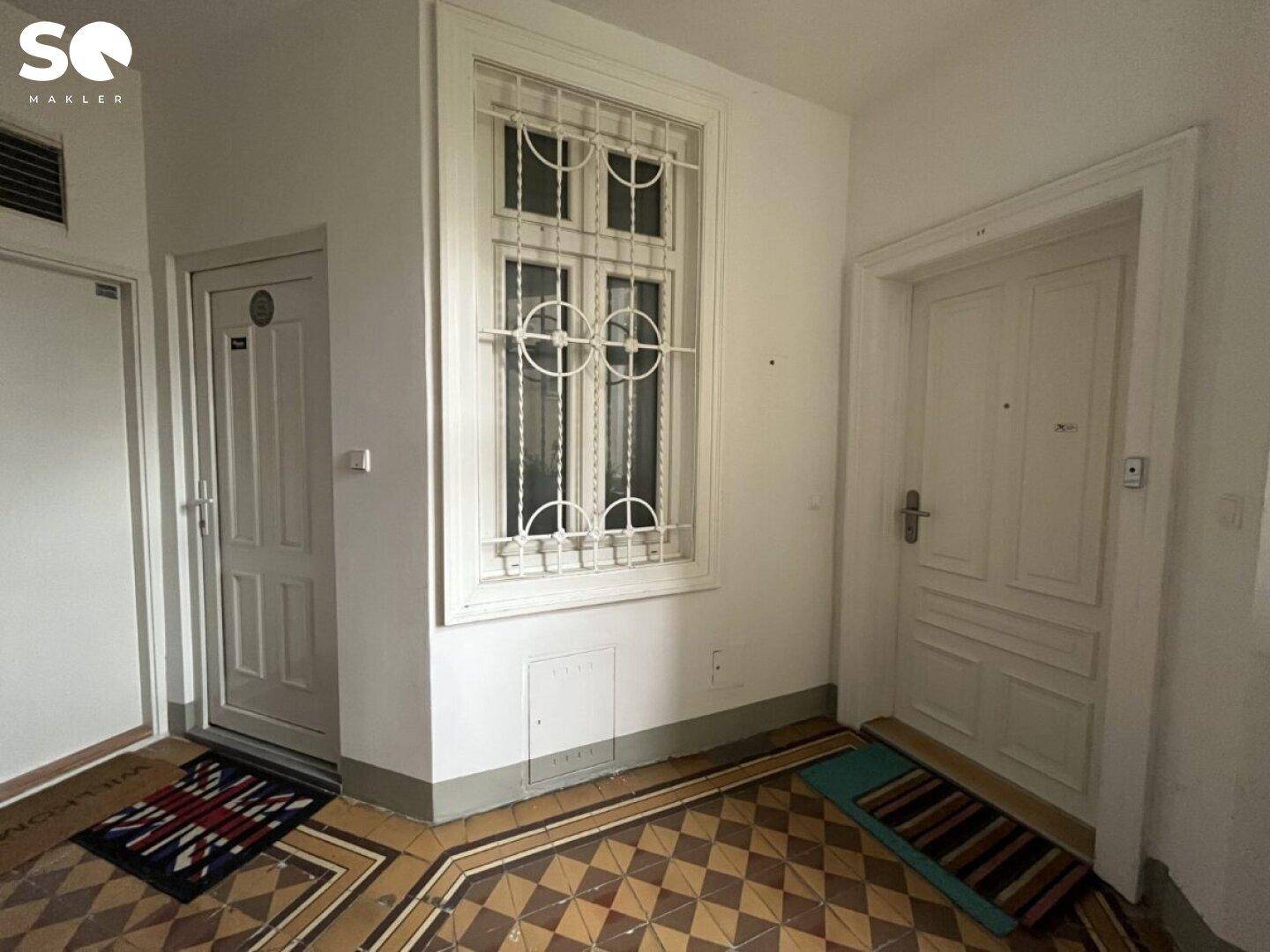 Eingangsbereich mit zwei Türen, gemustertem Fliesenboden und einem Fenster mit Gitter.