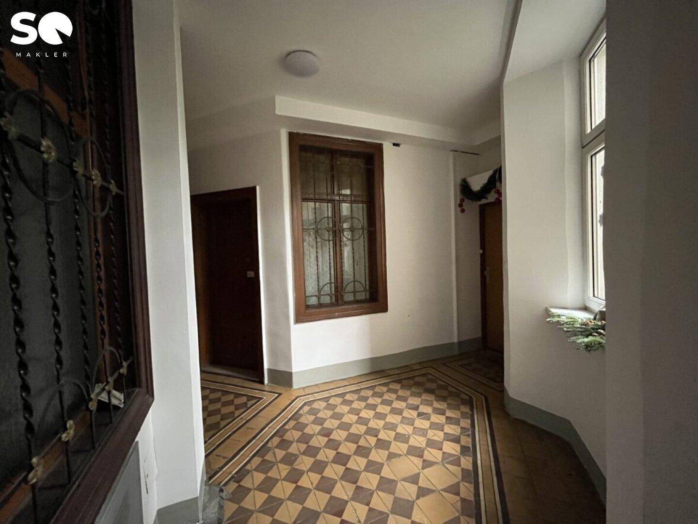 Historischer Flur mit gemustertem Fliesenboden und einem Fenster mit dekorativen Gittern.
