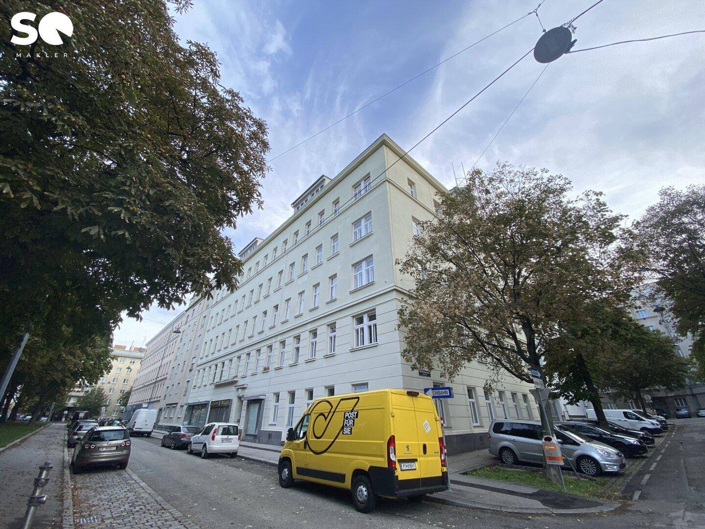 Mehrstöckiges Gebäude an einer von Bäumen gesäumten Straße mit parkenden Autos.