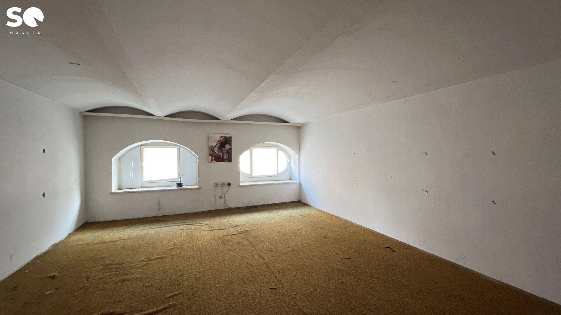 Leerer Raum mit gewölbter Decke, zwei Rundbogenfenstern und Teppichboden.