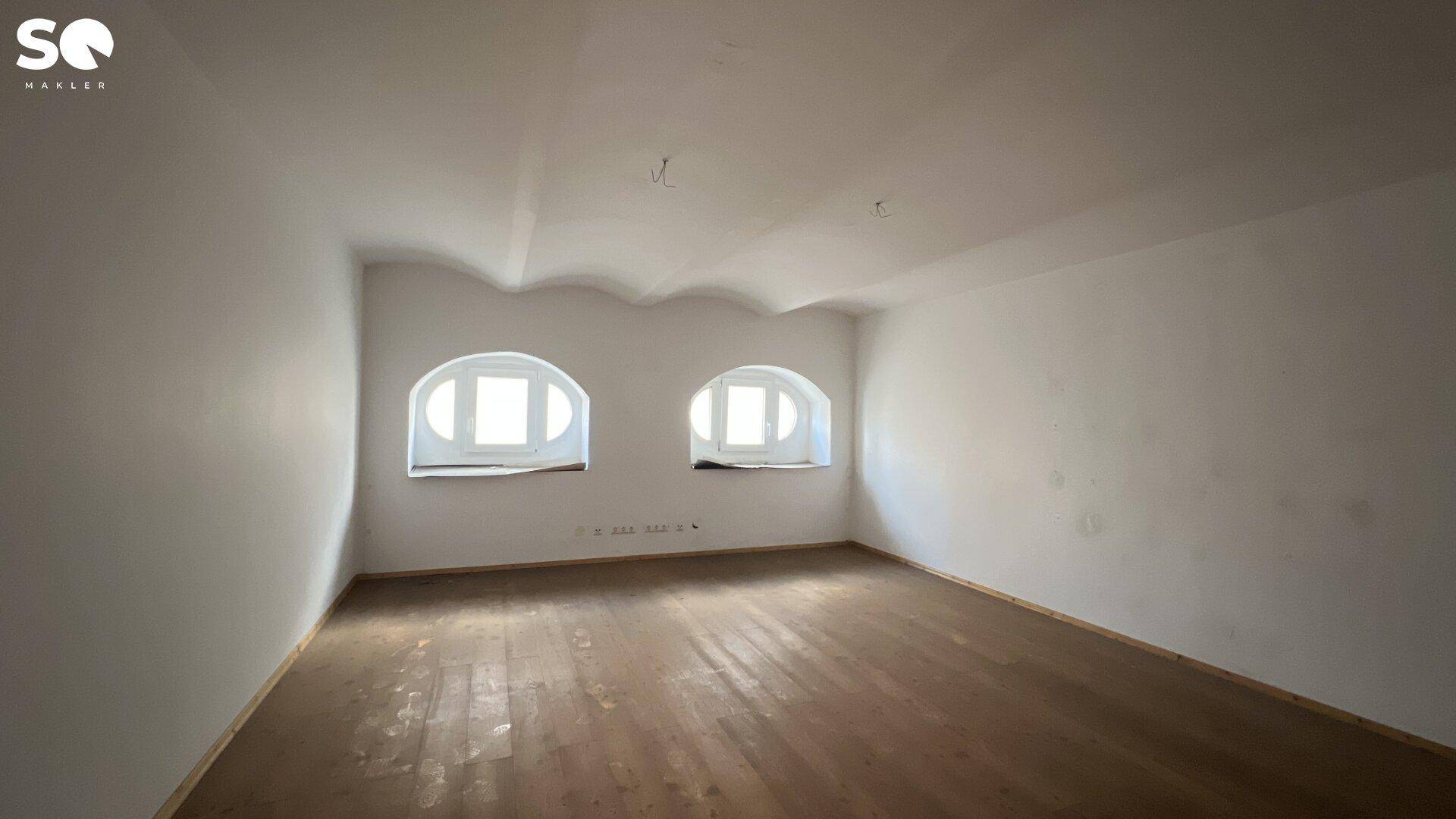 Leerer Raum mit Holzboden und zwei charakteristischen Rundbogenfenstern, hell und geräumig.