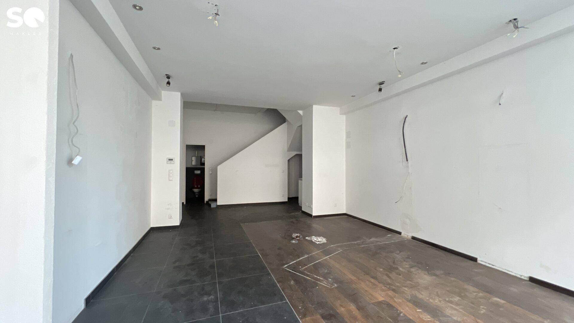 Geräumiges, leeres Ladenlokal mit unfertigem Boden und Zugang zu einem Treppenhaus.