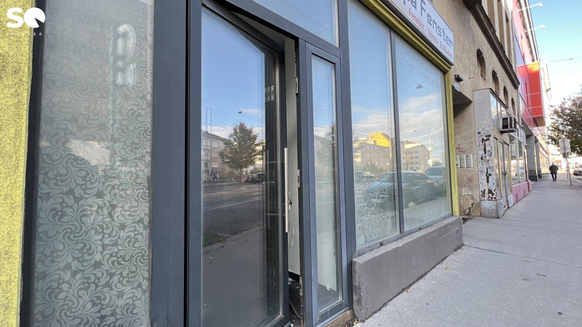 Eingangsbereich eines Geschäfts mit großen Schaufenstern an einer belebten Straße.