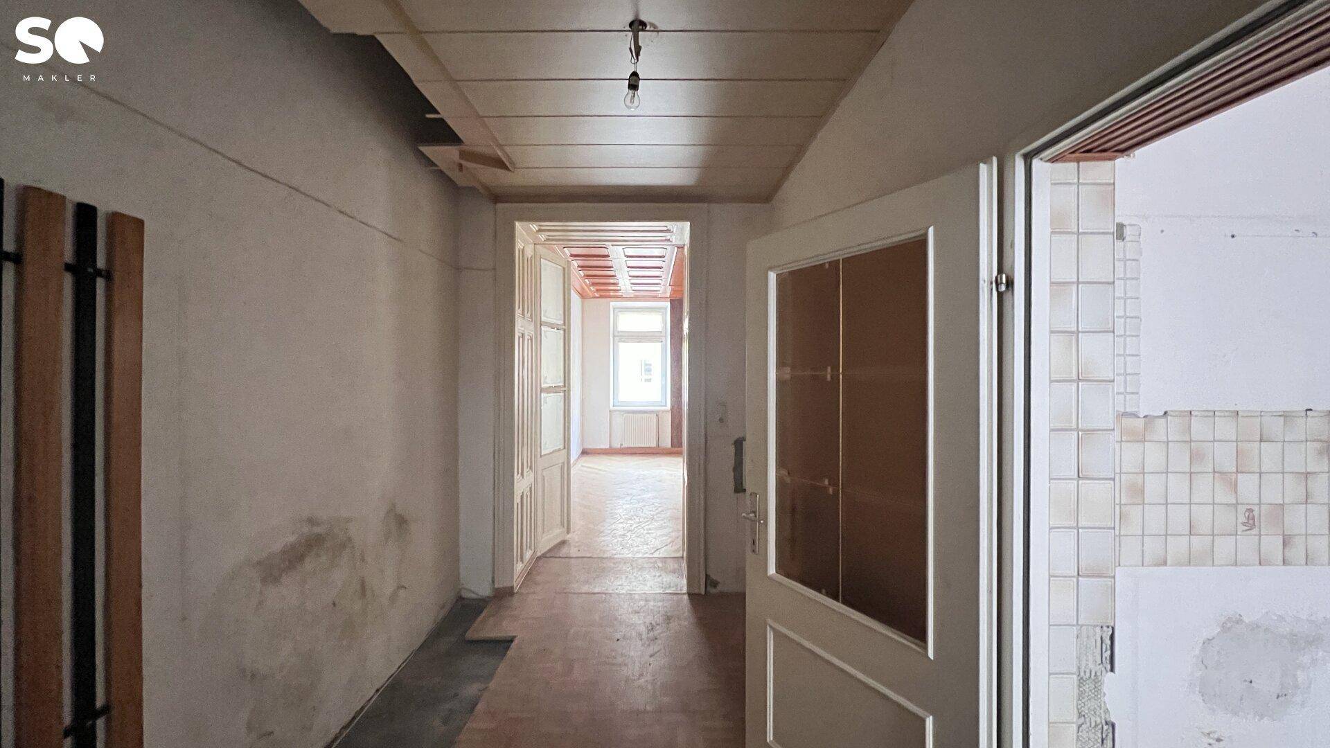 Renovierungsbedürftiger Flur mit unverputzten Wänden und einfacher Holzdecke, der zu weiteren Räumen führt.