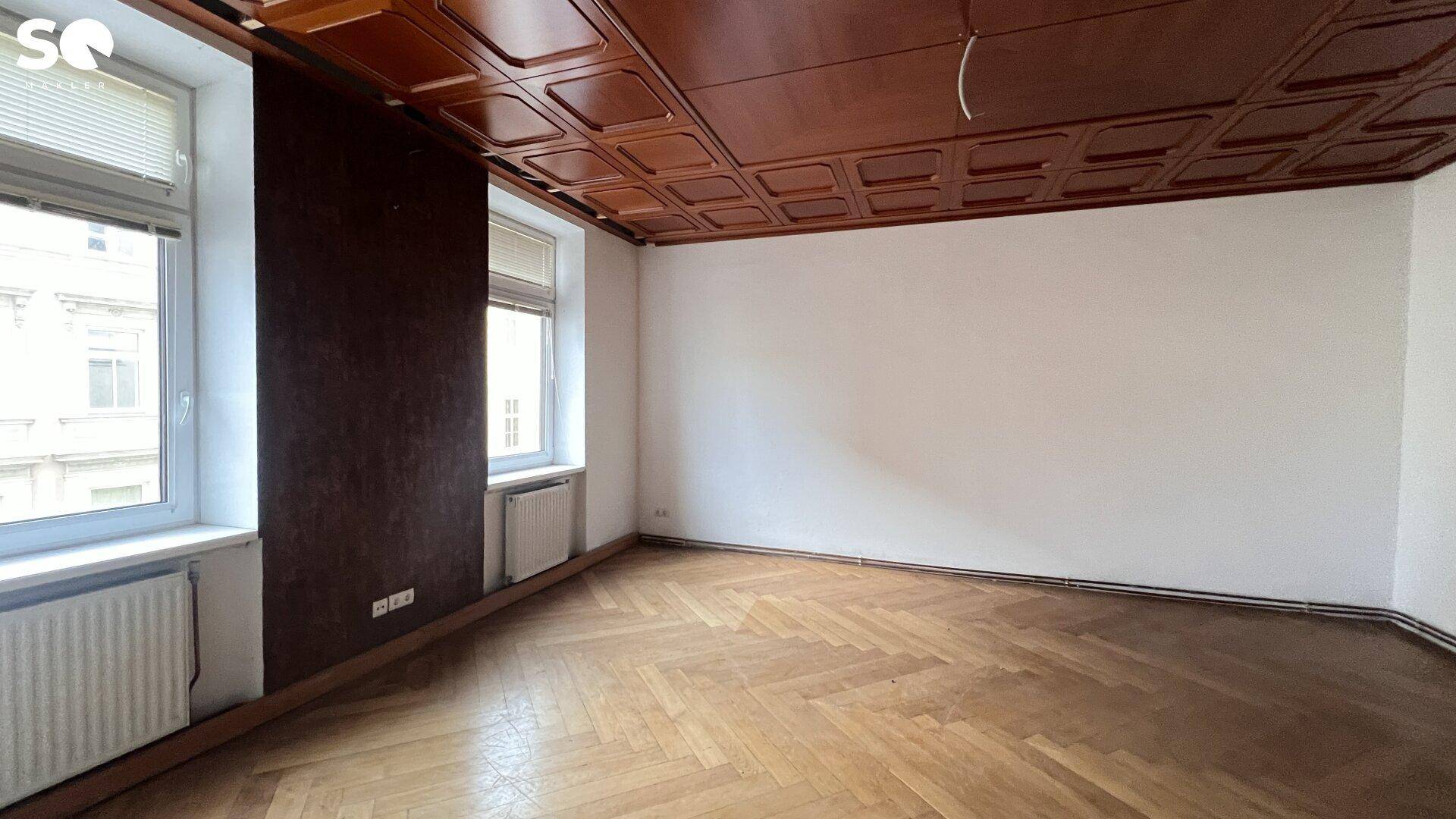 Leerer Raum mit Fischgrätparkett, weiß gestrichenen Wänden und einer dunklen Kassettendecke.