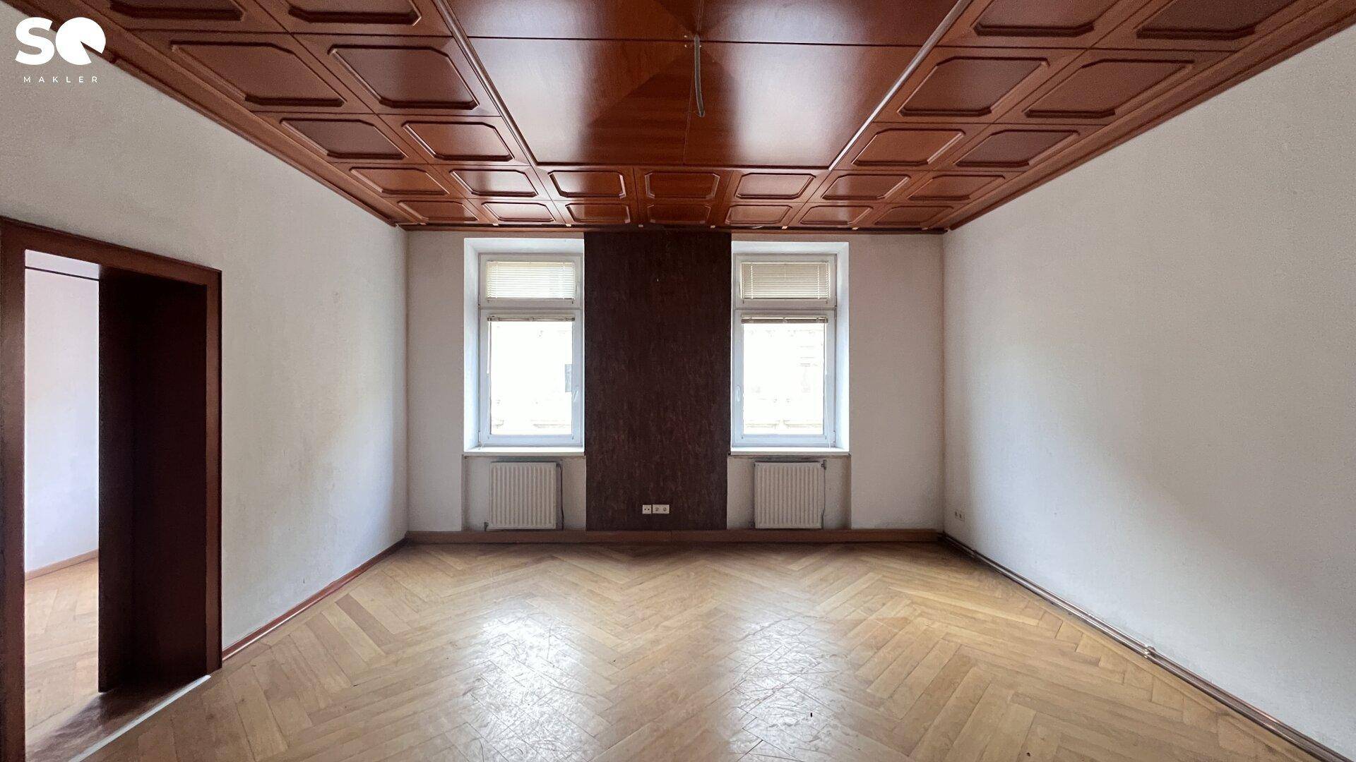 Leerer Raum mit Fischgrätparkett, weiß gestrichenen Wänden und einer dunklen Kassettendecke.