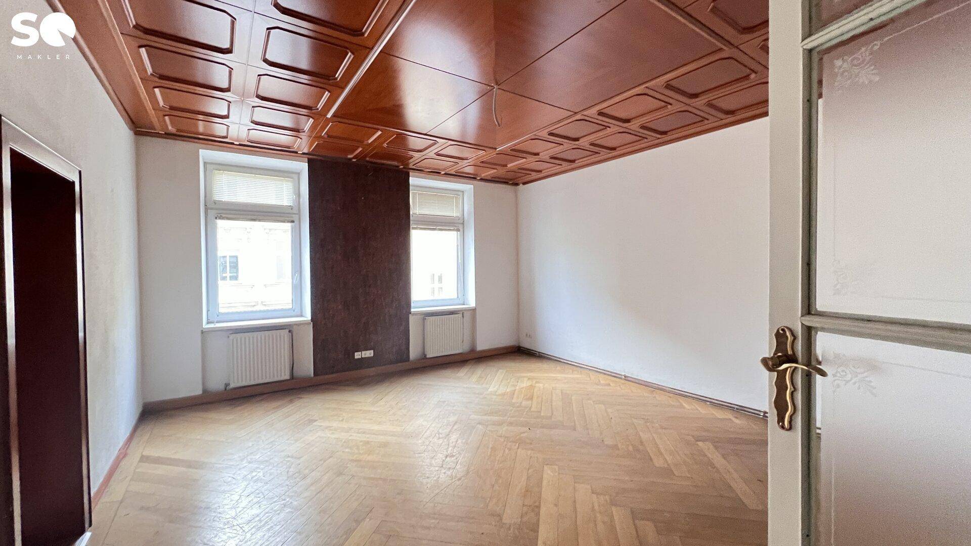 Leerer Raum mit Fischgrätparkett, weiß gestrichenen Wänden und einer dunklen Kassettendecke, vom Eingang aus gesehen.