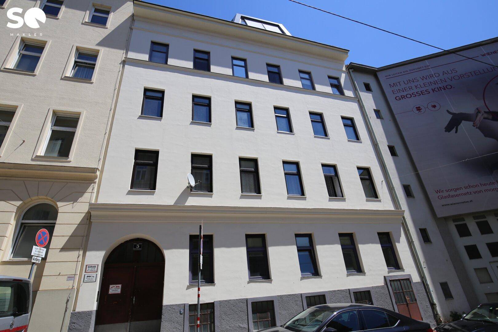 Weißes Gebäude mit dunklen Fenstern und geparkten Autos davor, an einem sonnigen Tag.