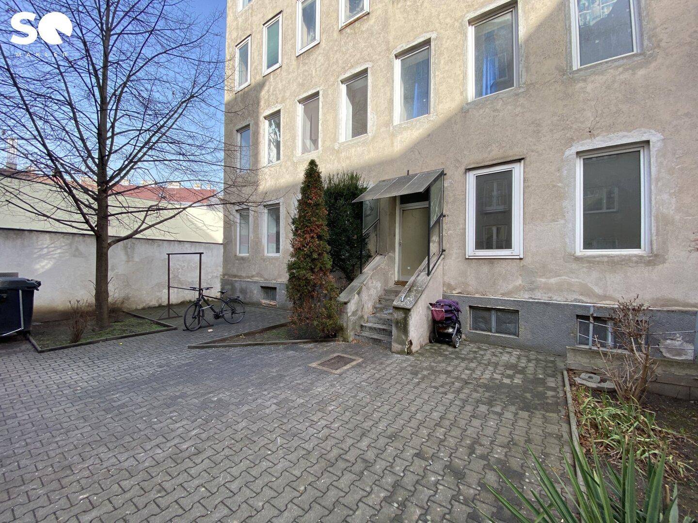 Eingangsbereich eines Mehrfamilienhauses mit Treppe, Vordach und gepflastertem Hof.
